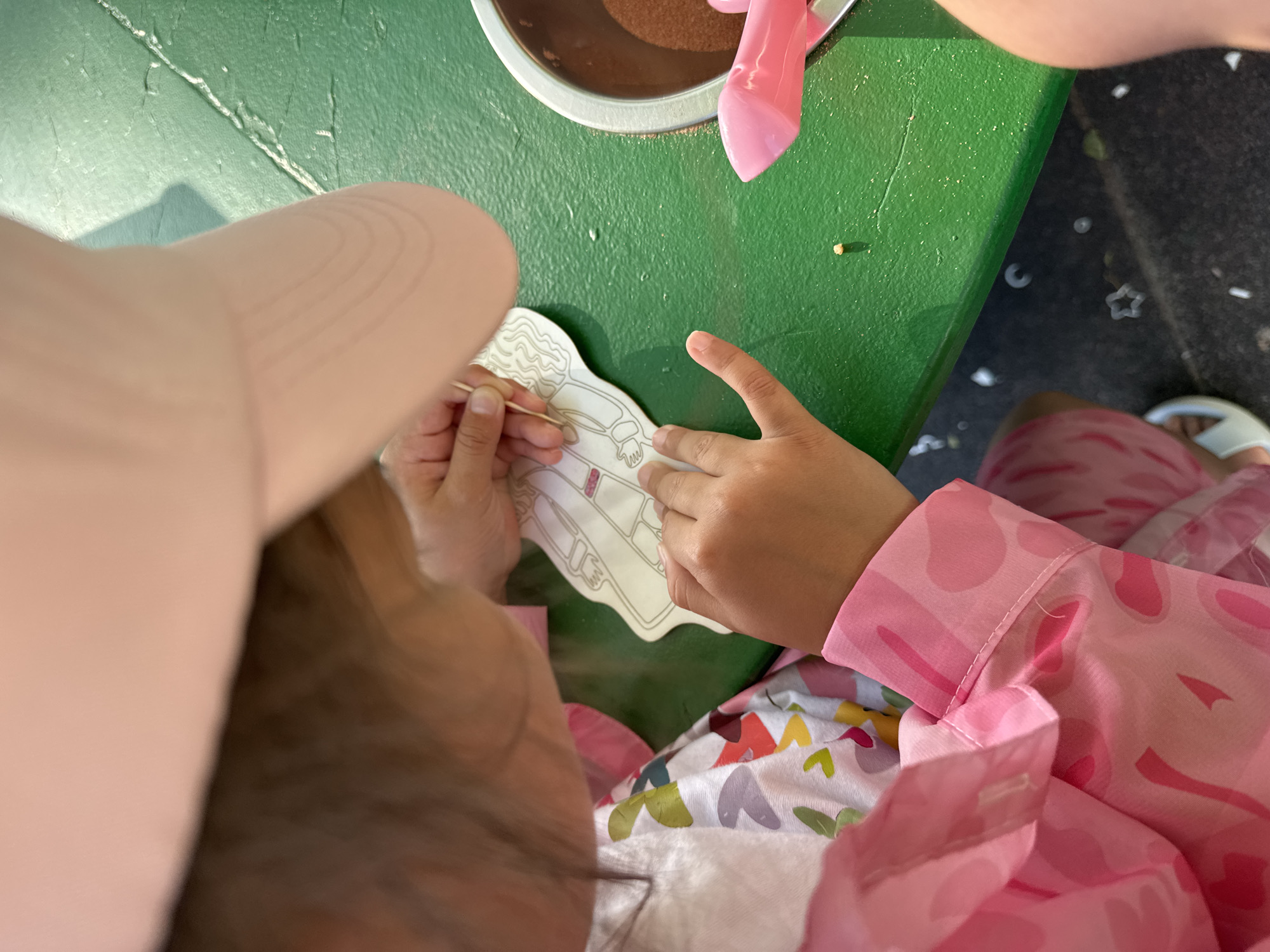 A forensic examination of the image reveals a top-down perspective of a young individual, likely a child, engaged in a crafting activity on a textured green surface, presumably a table, in Gelendzhik, Russia. The individual wears a light pink hat, a colorful patterned shirt with abstract shapes, and a pink long-sleeved smock featuring a subtle camouflage-like pattern. A pink cuff from the smock is visible on the right wrist. Parts of patterned pink lower garments and a white sandal are visible towards the bottom left.
The child's hands are central to the activity. The left hand stabilizes a white, die-cut paper outline of a figure, possibly a doll or character, with internal sections delineated. The right hand meticulously manipulates a thin wooden toothpick, appearing to apply or remove material from the paper figure's surface.
To the right of the activity, a metallic bowl with a silver rim contains a substantial quantity of a brown, granular material, consistent with sand or a similar craft medium. Adjacent to the bowl, a pink plastic tool, shaped like a small scoop or shovel, is partially visible. A small, light-colored speck rests directly on the green table surface near the center.
The green table exhibits minor surface irregularities and faint streaks. The background beneath the table is a darker, likely ground surface, upon which several small, white, star-shaped and crescent moon-shaped cutouts or debris are scattered. The scene is brightly illuminated, suggesting an outdoor or well-lit indoor environment.