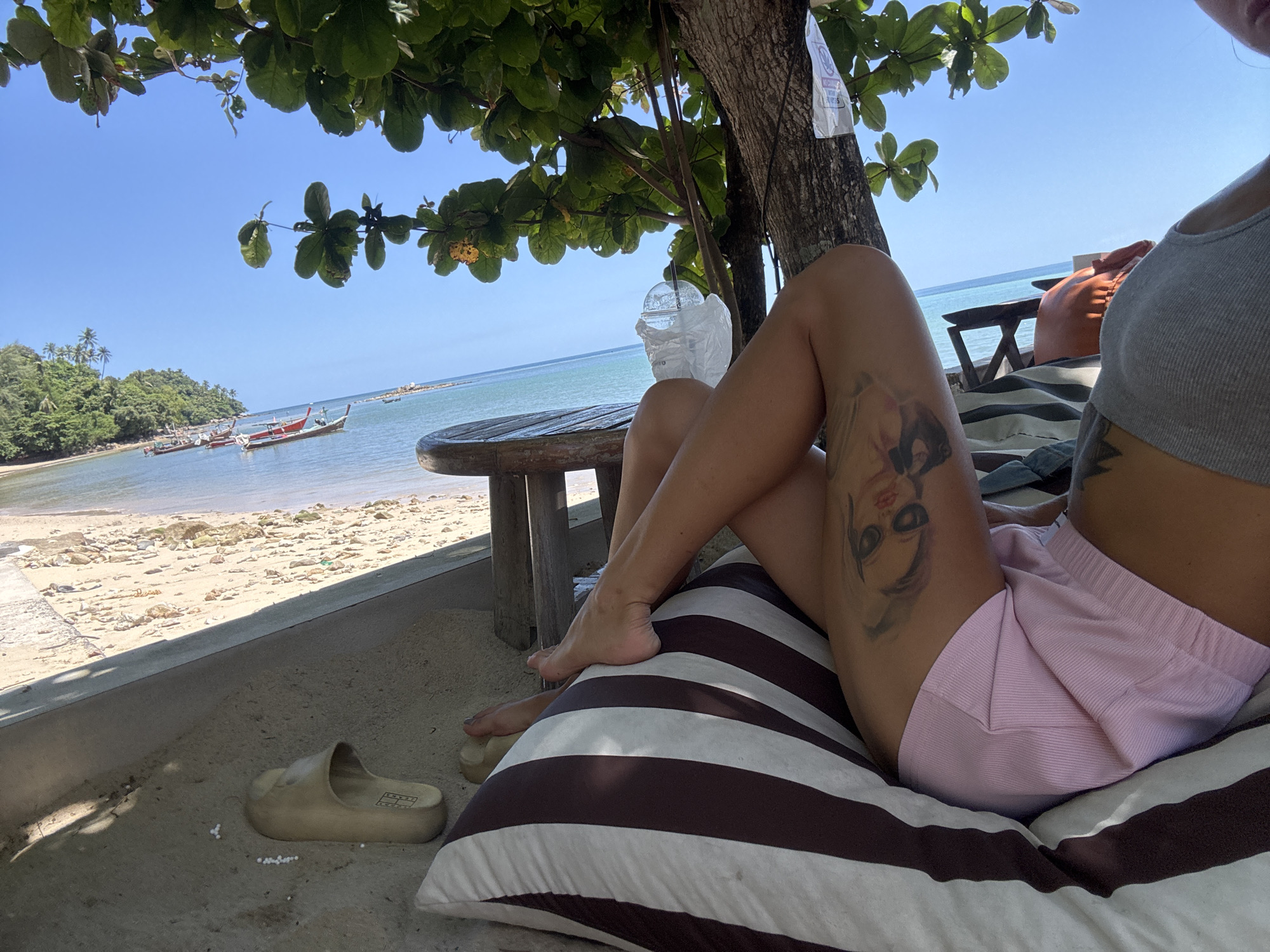A woman is relaxing on a striped cushion on a beach in Choeng Thale, Thailand. She is wearing a grey tank top and pink shorts, and has a tattoo on her thigh. Her feet are resting on the cushion. In front of her is a wooden table, and beyond that, the beach and the ocean. There are several longtail boats in the water, and palm trees on the shore. The sky is clear and blue. A light-colored slide is on the ground next to the cushion.