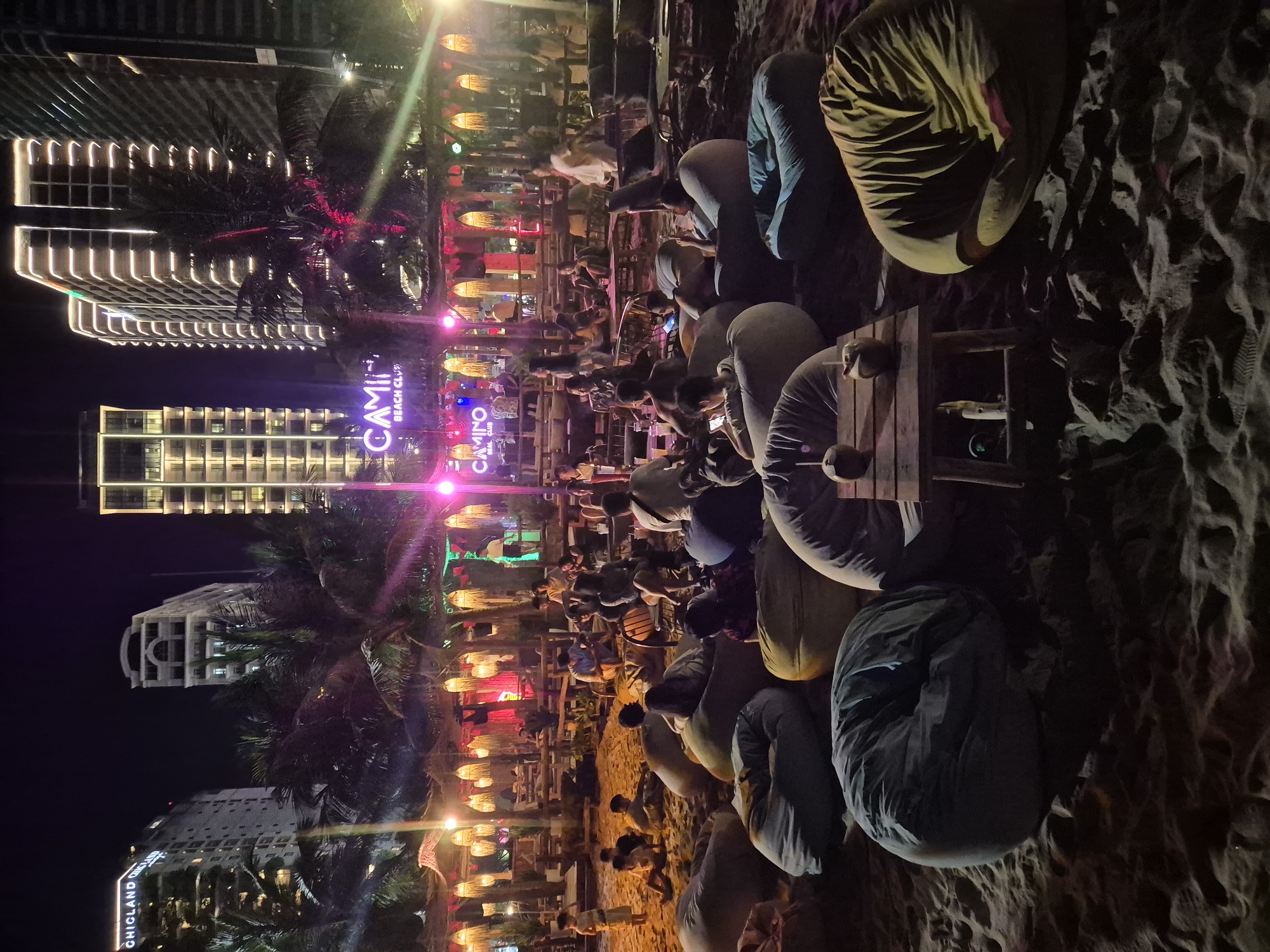 This image depicts a nighttime scene at "Camino Beach Club" located in the city of Đà Nẵng, Vietnam. The beach club is situated on a sandy area, with a stage in the background featuring a sign that reads "CAMINO BEACH CLUB." The venue is illuminated by numerous decorative lights, including string lights and spotlights, casting a vibrant ambiance. In the foreground, several large beanbag chairs are scattered across the sand, occupied by individuals of various ages. A small wooden table with two coconut drinks is placed in front of some of the beanbags. In the middle ground, more people are seated at tables and on low seating arrangements, some appearing to be patrons of the establishment, while others might be staff. The backdrop reveals several tall, modern buildings, characteristic of a coastal city's skyline. Palm trees are also visible, adding to the tropical beach setting. The overall scene suggests a lively and social gathering at a beachside establishment.