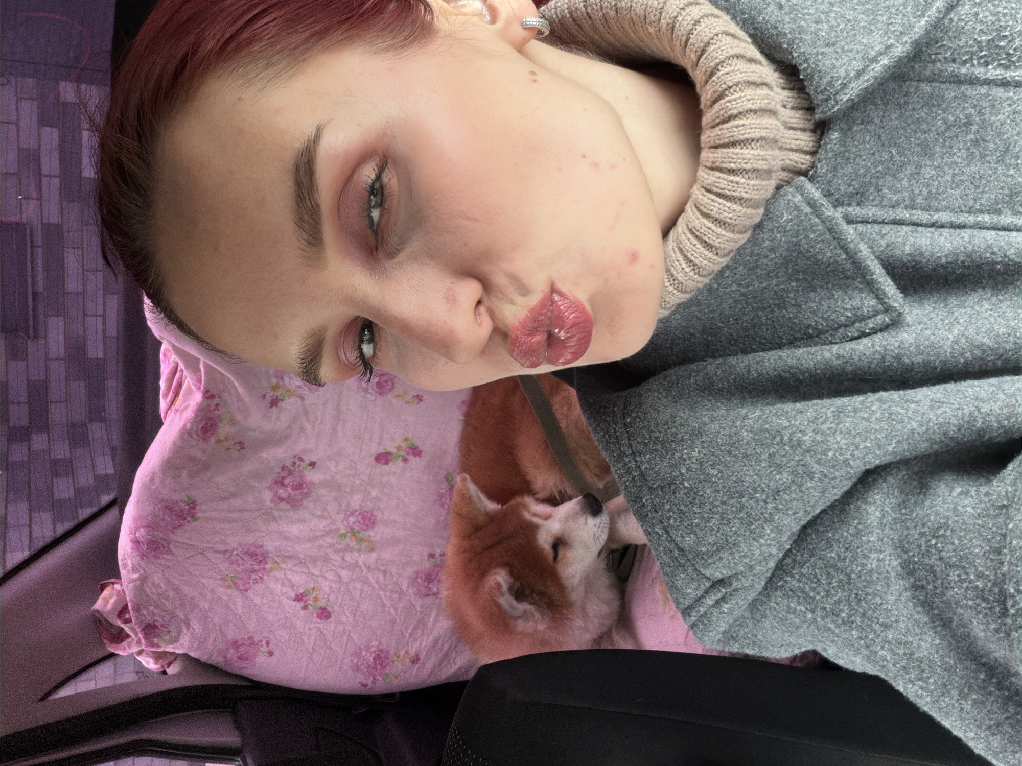 The image captures a close-up, tilted view of a woman and her dog resting inside what appears to be a car, possibly in Chaikovsky, Russia. The woman, with red hair and light skin, is positioned with her head tilted, making a playful "kissing face" towards the camera, her lips pursed and glossy. She has green eyes, prominent eyebrows, and is wearing visible eye makeup. Her attire includes a gray, possibly wool, coat over a ribbed beige turtleneck or sweater. A silver hoop earring is visible in her right ear. Some minor skin blemishes are present on her face.
Next to the woman, nestled on a soft, pink quilted pillow with a floral (rose) pattern, is a reddish-brown and white dog, resembling an Akita or Shiba Inu. The dog appears to be resting peacefully, with its head turned slightly away from the camera.
The background suggests an enclosed space, with parts of a black car seat visible. Through a window, a blurry purple-hued structure, possibly a brick or tiled wall, is visible, indicating an outdoor urban or built-up environment. The overall scene conveys a sense of relaxed companionship during travel. There is no visible text in the image.