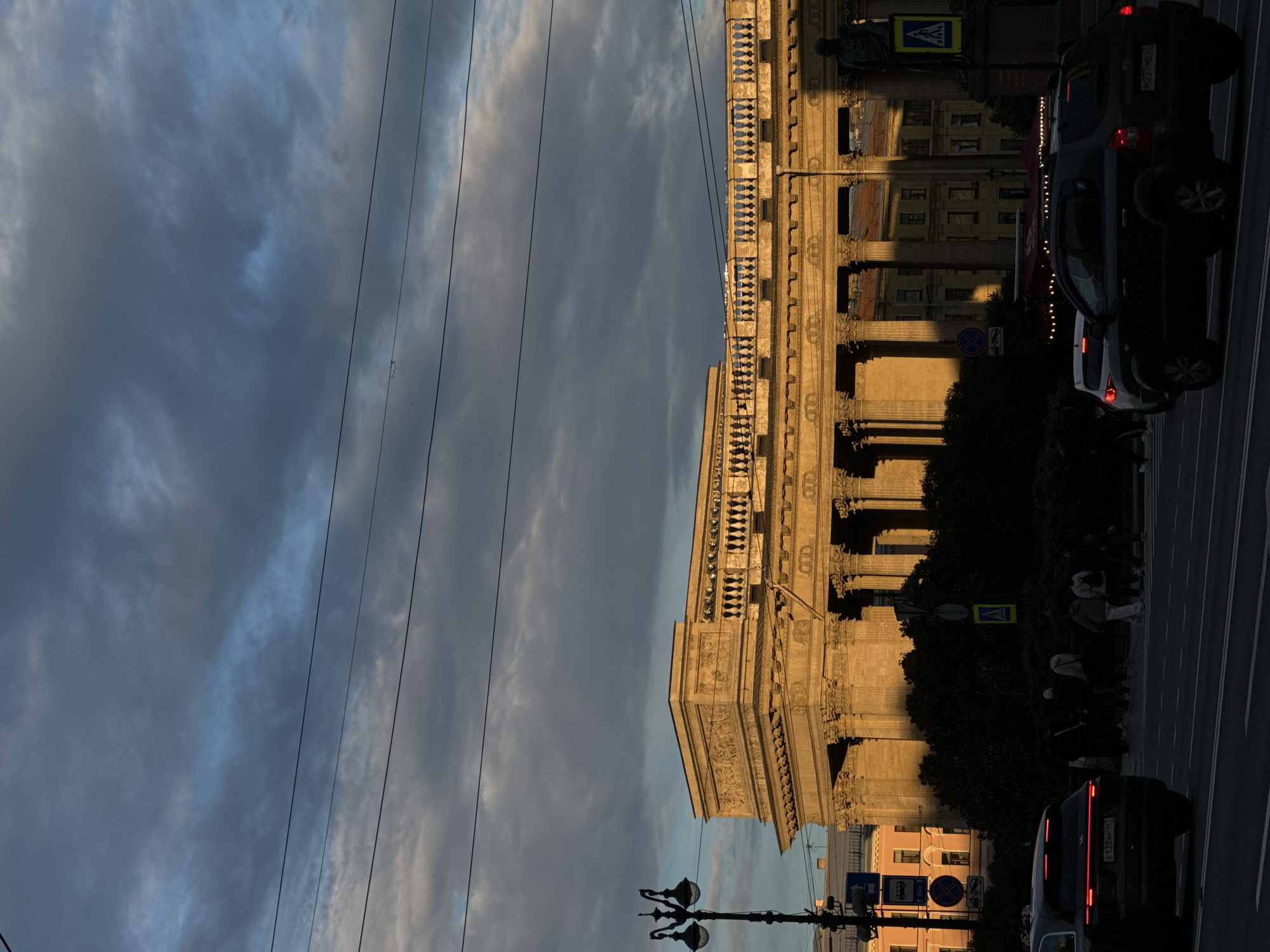 The image depicts Kazan Cathedral in Saint Petersburg, Russia, during a late afternoon or early evening with dramatic lighting. The facade of the cathedral, characterized by its grand colonnade and neoclassical architecture, is bathed in the warm, golden light of the setting sun. The sky above is filled with heavy, dark clouds, interspersed with patches of lighter blue, creating a stark contrast with the illuminated building. Several power lines crisscross the upper portion of the frame.
In the foreground, a multi-lane road is visible, with several vehicles driving or stopped. On the left, the rear of a dark-colored SUV with illuminated red taillights is prominent. To the right, another dark SUV is in motion. Pedestrians are crossing the street at a marked crosswalk, their figures silhouetted against the bright facade of the cathedral. They are a mix of adults and possibly children, engaged in the act of crossing.
A vintage-style lamppost stands on the left side of the image, near the road. Street signs, including traffic signals and pedestrian crossing symbols, are also visible. The surrounding environment includes trees and other buildings, some of which are partially obscured by the cathedral's structure and the shadows cast by the low sun. The overall scene conveys a sense of urban life unfolding under dynamic atmospheric conditions.