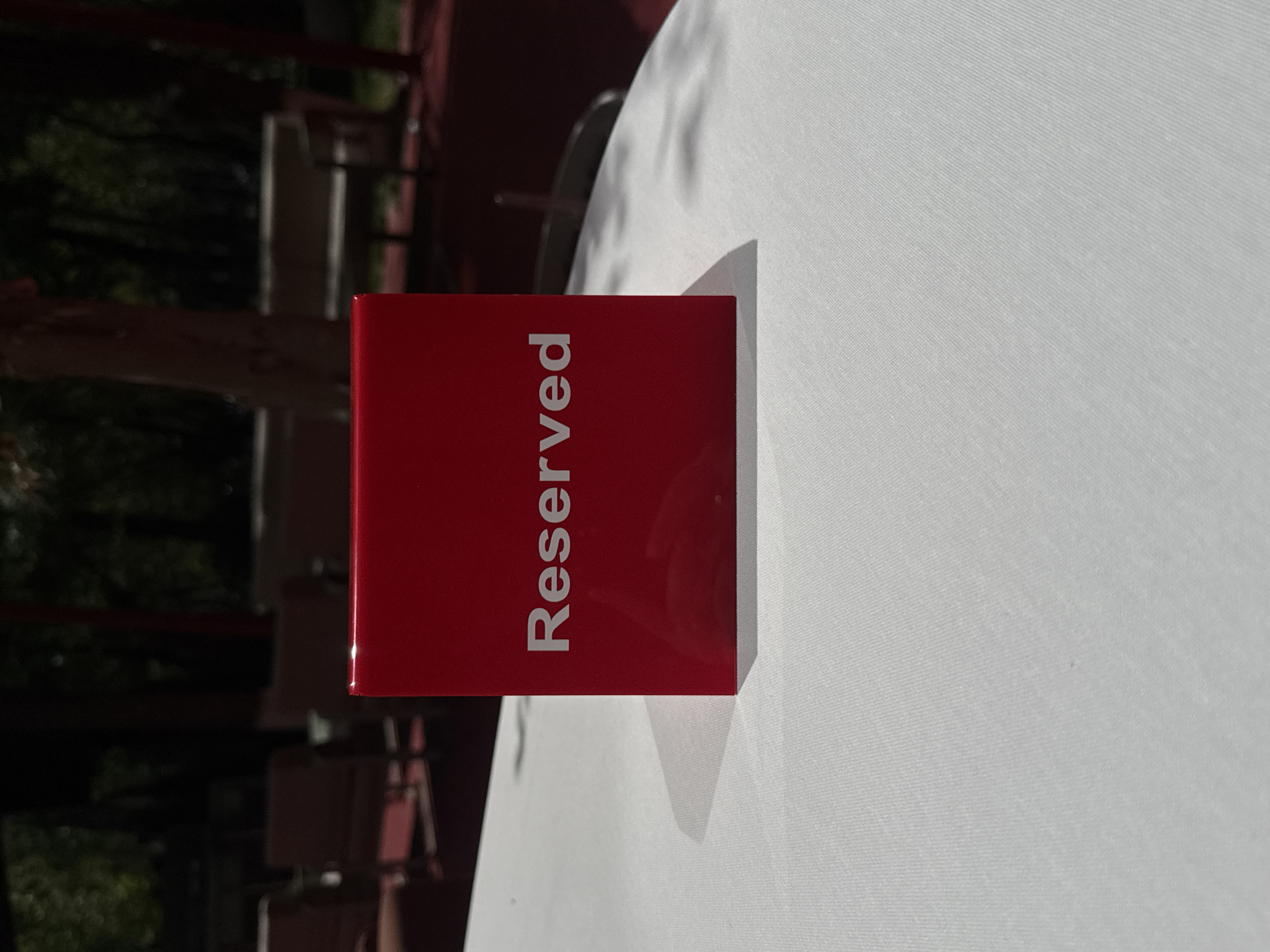 A red, rectangular sign, displaying the word "Reserved" in white sans-serif letters, is centrally featured on a pristine white tablecloth. The image is rotated 90 degrees clockwise, causing the text to appear vertically oriented. Long, distinct shadows are cast from the sign onto the tablecloth, indicating bright natural lighting, likely during the day. The background is softly blurred, revealing dark, indistinct shapes that suggest trees or other outdoor elements, along with some dark structures, possibly furniture, in what appears to be an outdoor dining area or a restaurant with a view. This scene depicts a table set aside for future patrons in Yerevan, Armenia.