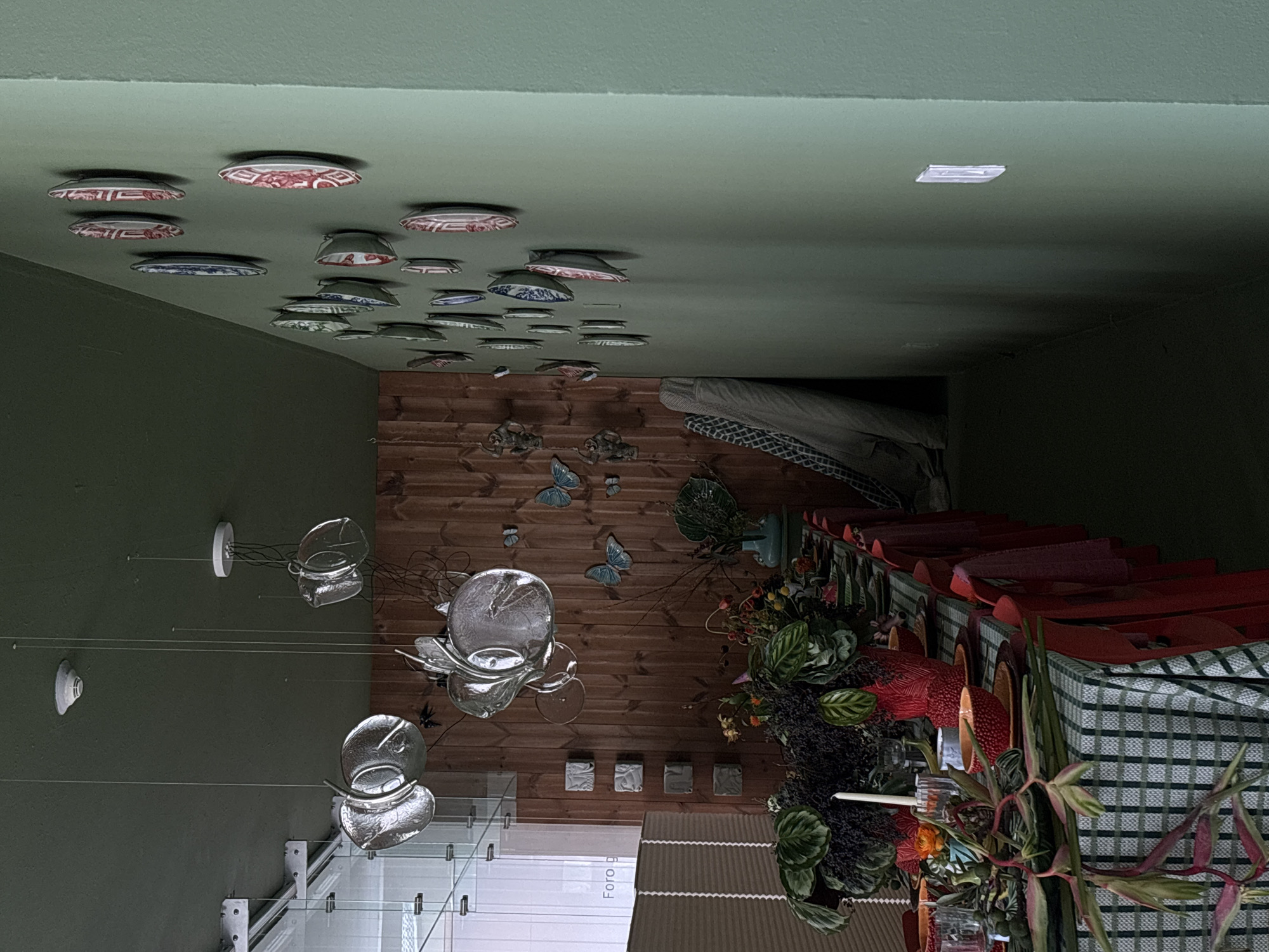 This image, likely taken indoors, reveals a room with a green ceiling adorned with numerous decorative plates. A multi-pendant light fixture hangs on the left side of the frame. A wooden wall decorated with butterfly figures is visible. The right side of the image shows a table setting with plants and a checkered tablecloth. The location is inferred to be Mexico City, Mexico, though there is no visual confirmation of this information in the image. No people are visible in the frame.