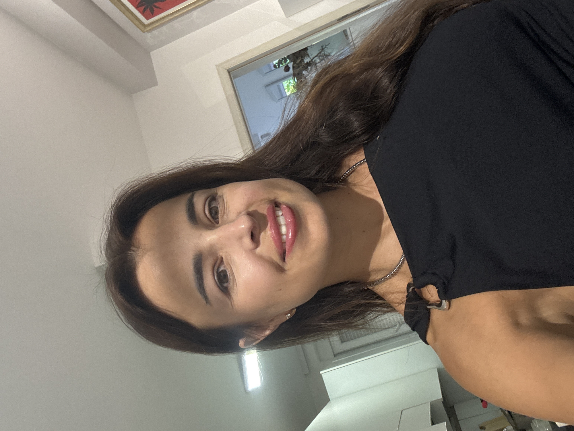A woman with long, dark brown hair and brown eyes poses for a selfie in what appears to be a kitchen. She is wearing a black top with a silver necklace. Her skin is lightly tanned, and she is smiling. The background shows kitchen cabinets and a doorway leading to another room. There is a framed picture on the wall to the right. The lighting suggests an indoor environment, possibly at night.