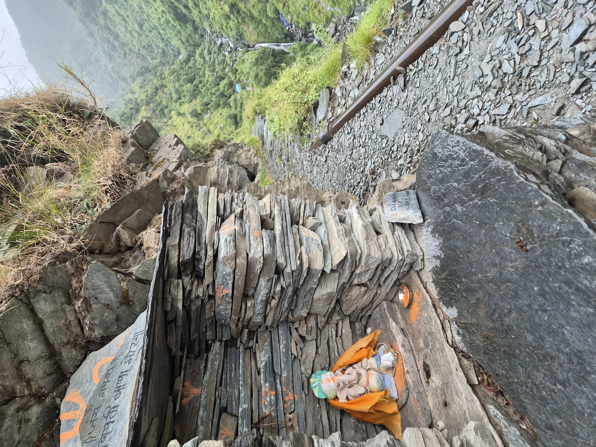 In Bhagsu, India, a small shrine is nestled into a rocky overhang. The shrine is constructed of stacked slate stones and houses a small idol of the Hindu deity Ganesha, adorned with an orange cloth. In front of the idol, a small dish holds an orange liquid, possibly oil for a lamp, and a small lit diya is visible. Orange markings, possibly religious symbols, are painted on some of the slate stones. To the right of the shrine, a stone pathway leads into a lush green valley with a waterfall cascading down. A metal pipe runs alongside the path. A small sign on a rock reads "CLEANER & GREENER". The overall environment suggests a remote, natural setting.