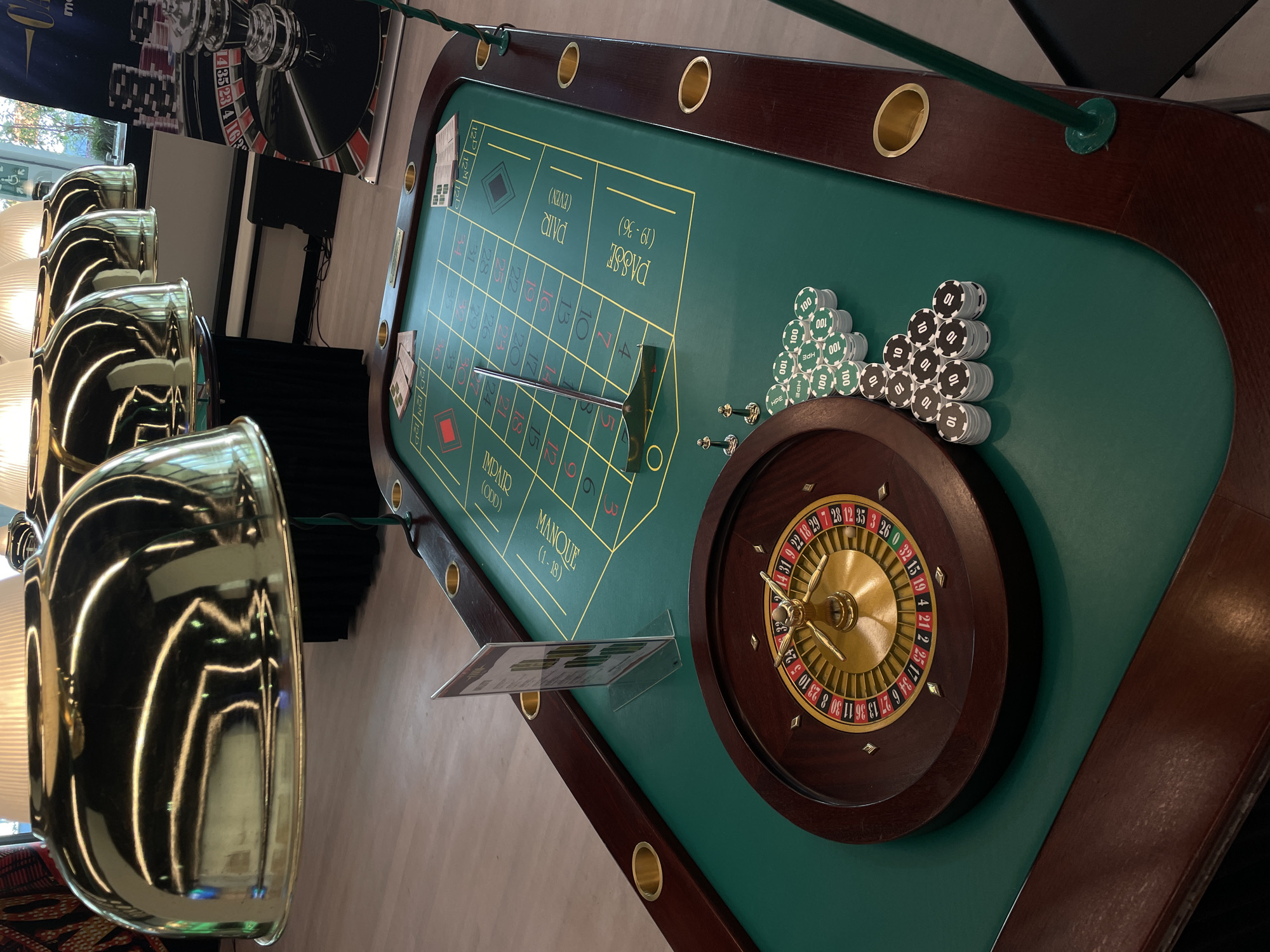 This image shows a close-up, high-angle shot of a roulette table. The table is green with a dark wood trim. The roulette wheel is positioned on the right side of the table, and stacks of casino chips are located beside it. A silver rake rests on the table's surface near the numbered grid. The table's layout includes sections labeled with terms like "PAIR (EVEN)", "PASSE (19-36)", "MANQUE (1-18)", and "IMPAIR (ODD)". Gold-colored light fixtures are seen in the background on the left, and a banner with a roulette wheel image is visible in the back.