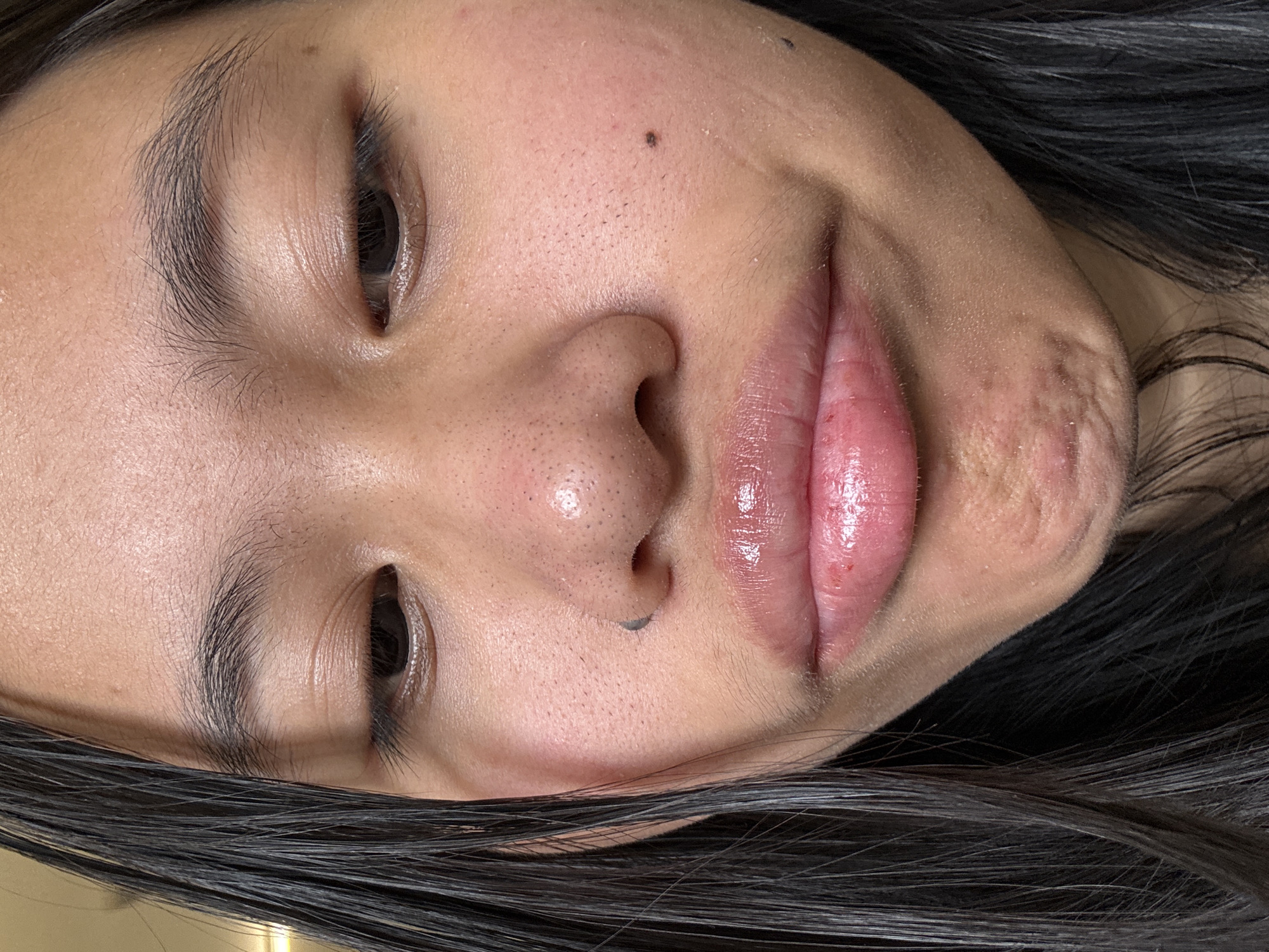 A close-up, waist-up shot features a young woman looking down, her face angled slightly downwards and to the right. Her dark hair is styled down, with strands falling over the left side of her face. She has light brown eyes, arched eyebrows, and a clear complexion with some visible pores on her nose and cheeks. Her lips are full and shiny, with a glossy pink lipstick. There is a small dark mole on her left cheek and another on her nose. The background is blurred, suggesting an indoor environment, possibly a home or studio. The lighting is soft, casting a warm glow on her face. The image appears to be taken in East Jakarta, Indonesia.