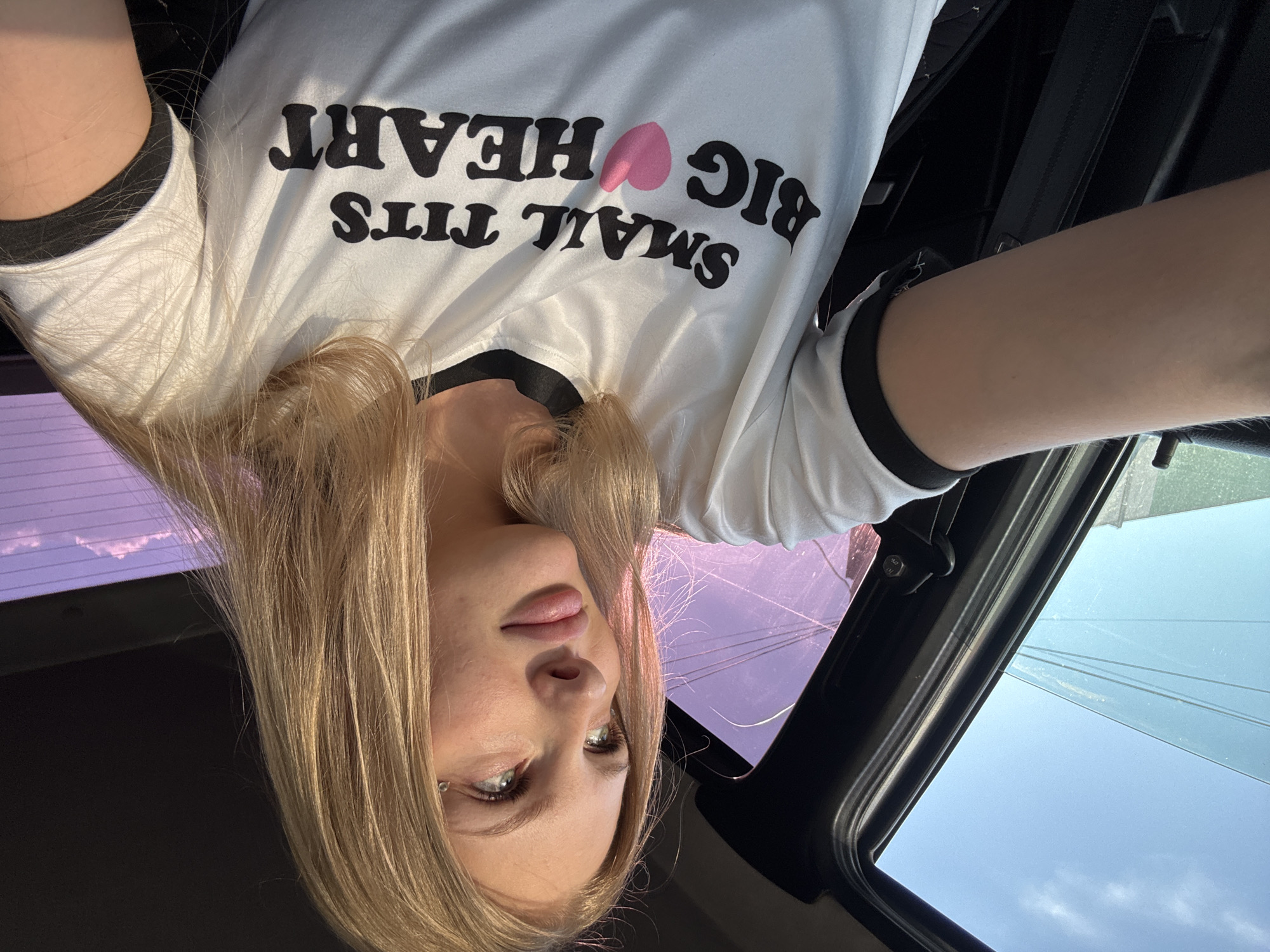 The image shows a young woman with blonde hair and light skin sitting in the passenger seat of a vehicle. She is wearing a white t-shirt with black trim and a black inscription that reads "SMALL TITS BIG HEART" with a pink heart symbol. The woman is looking out the window to her right, her gaze directed upwards. The interior of the car is visible, including a seatbelt and parts of the car door. Outside the window, the sky transitions from a clear blue to shades of purple and pink, suggesting it is either sunrise or sunset. The background shows some indistinct structures and power lines. The image appears to be a selfie taken from the perspective of someone in the driver's seat. There is no visible indication of the specific location being the city of Perm, Russia.