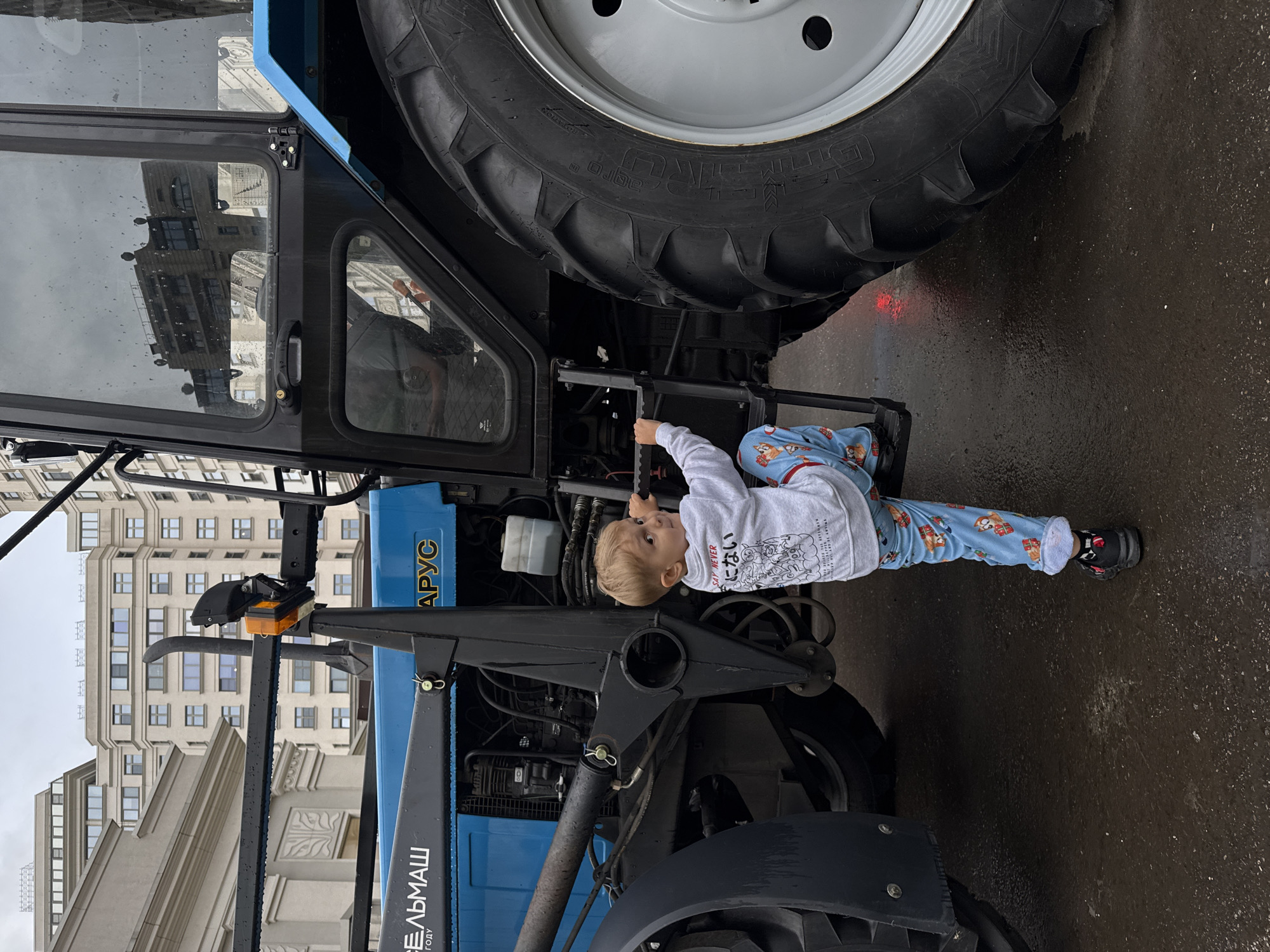A young boy, appearing to be around 3-4 years old, is climbing the steps of a large blue tractor. He is wearing a grey sweatshirt with Japanese text and an illustration, and blue pants with a pattern of what appear to be cartoon animals and gifts. He is looking to his right, with his right hand gripping a metal bar. The tractor is large with a prominent black tire and a blue cab. The scene is set in an urban environment, with tall buildings visible in the background. The ground is wet, suggesting it may have recently rained. The overall impression is one of a child exploring a large piece of machinery in a city setting.