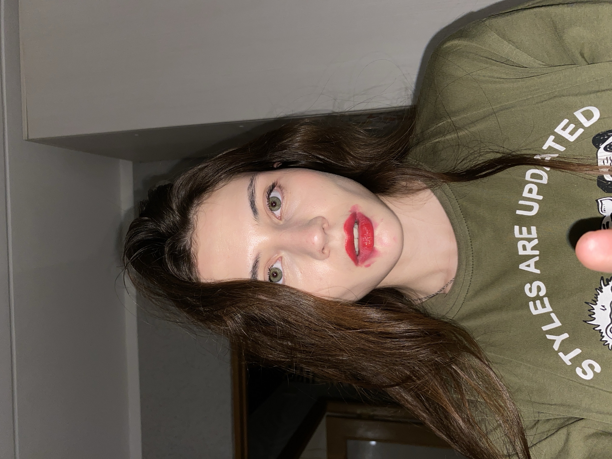 A close-up portrait shows a young woman with long, wavy dark brown hair, oriented as if lying down and looking directly at the viewer. Her striking green eyes are open wide. Her lips are adorned with bright red lipstick, which is noticeably smudged around her mouth, suggesting either a playful act or recent activity. Her skin appears fair with some highlights on her forehead and cheeks. She is wearing an olive green t-shirt with white text, part of which reads "STYLES ARE UPDATED." A partial graphic, possibly a cartoon character like a panda, is visible below the text on her shirt. The background is dimly lit and indistinct, suggesting an indoor setting with elements like a light-colored wall or cabinet above her head and a darker wooden surface or furniture in the lower right. The overall lighting seems to be artificial. Her expression is neutral, engaging directly with the camera.