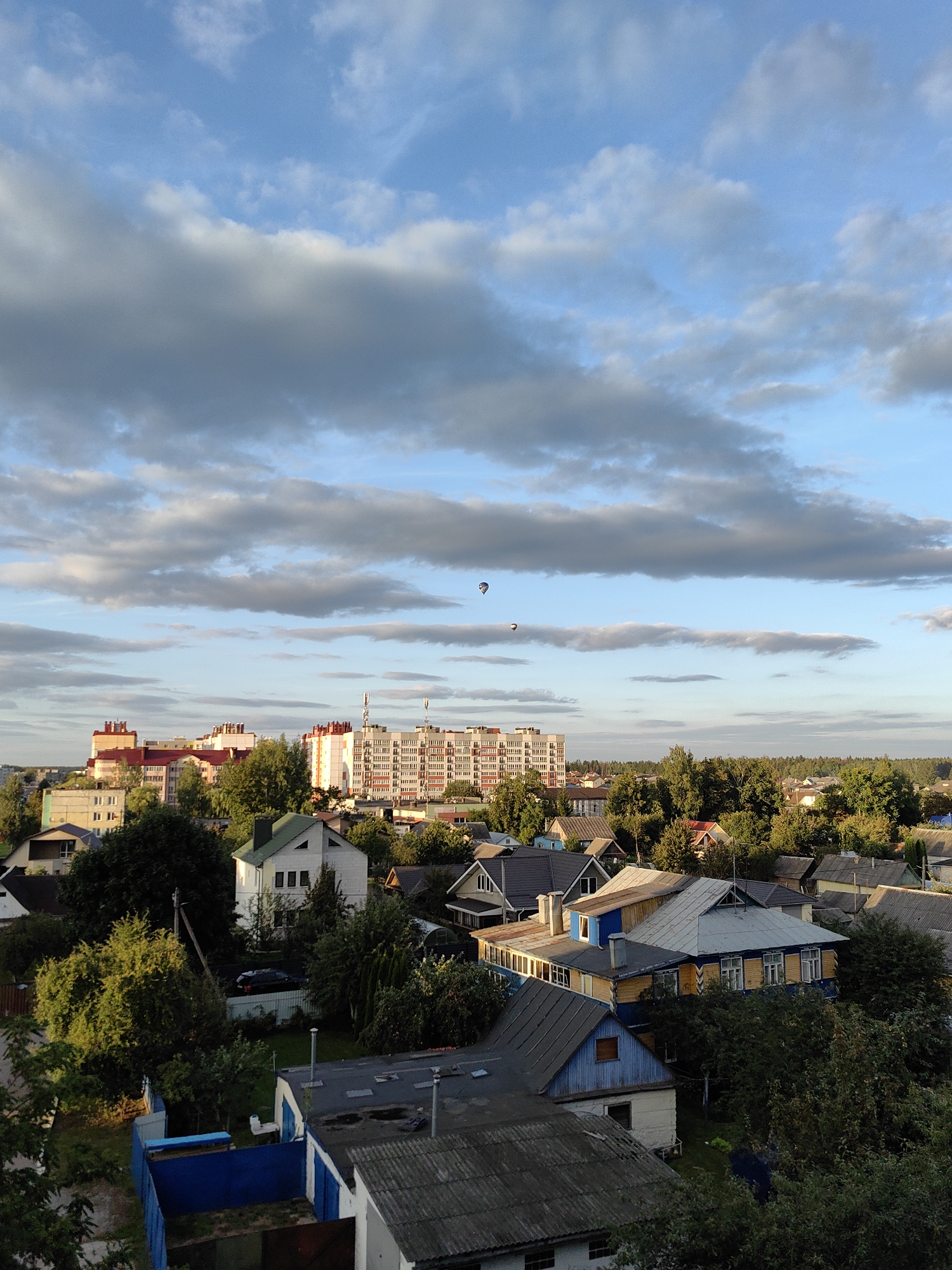 An aerial, high-angle, full shot captures a sunlit residential area in Fanipal, Belarus, under a sky filled with scattered cumulus clouds. In the foreground, a cluster of rooftops of various architectural styles dominate the scene, with some featuring weathered grey shingles and others with brighter, more modern materials. Many of these homes are nestled among lush green trees, suggesting a well-established neighborhood.
In the mid-ground, a large, multi-story apartment building stands out with its light-colored facade and numerous windows, indicating a more urban element within the otherwise suburban landscape. The apartment building is surrounded by more trees and smaller residential structures.
The sky above is a vibrant blue, punctuated by dramatic, elongated clouds that cast soft shadows over the town. In the distance, a dense forest line marks the edge of the developed area. Two hot air balloons are visible in the sky, appearing as small, dark specks against the clouds and blue expanse, adding a touch of aerial activity to the scene. The lighting suggests it is either early morning or late afternoon, with the sun casting a warm glow on the buildings and foliage.