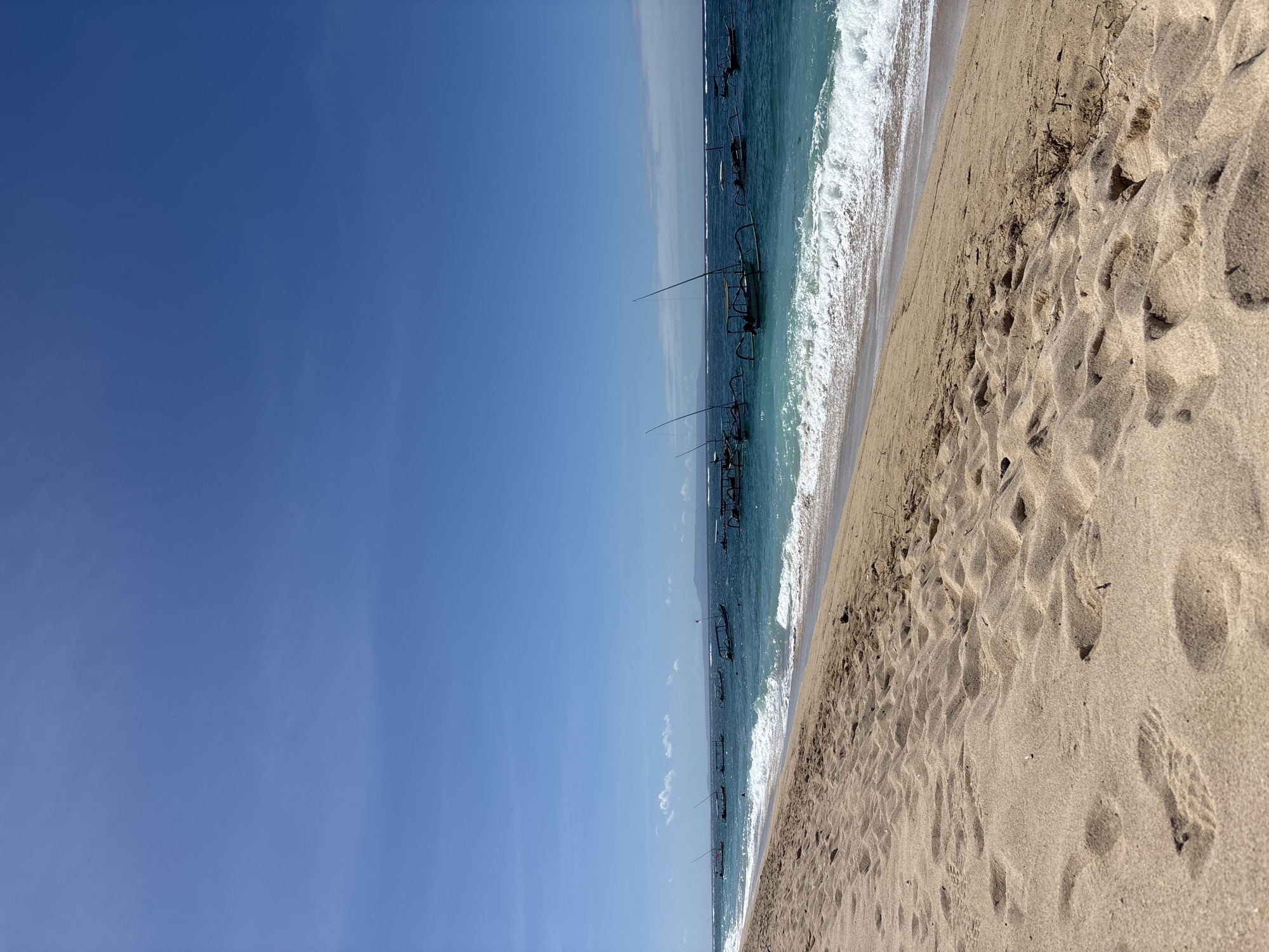 The image depicts a beach scene in Kuta Selatan, Indonesia. The sky is clear blue. The ocean, with varying shades of blue, has waves washing toward the shore. Several boats are visible in the water, aligned parallel to the shore. The beach is sandy, with footprints.