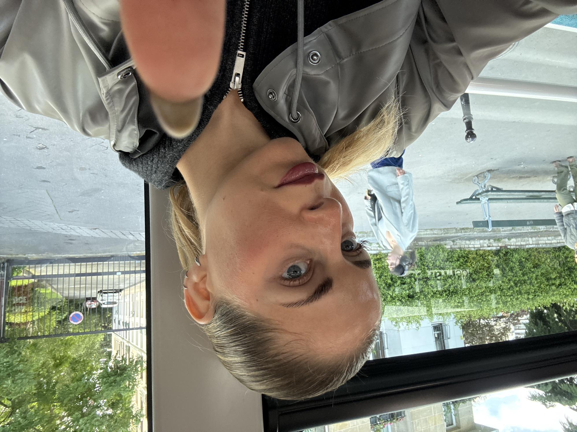A selfie photo taken by a woman on a public transport vehicle, possibly a bus or tram. The woman has light skin, blonde hair slicked back, and is wearing a grey jacket over a black sweater. A blurred thumb obstructs the upper left corner of the image. Through the window behind her, a street scene is visible. There's a metal gate with a "SENS UNIQUE" sign (meaning "one way" in French). Further in the background, there are buildings and trees. A couple of people can be partially seen in the reflection on the window. The scene implies an urban environment, with a possible location in Paris, France based on the road sign in French.