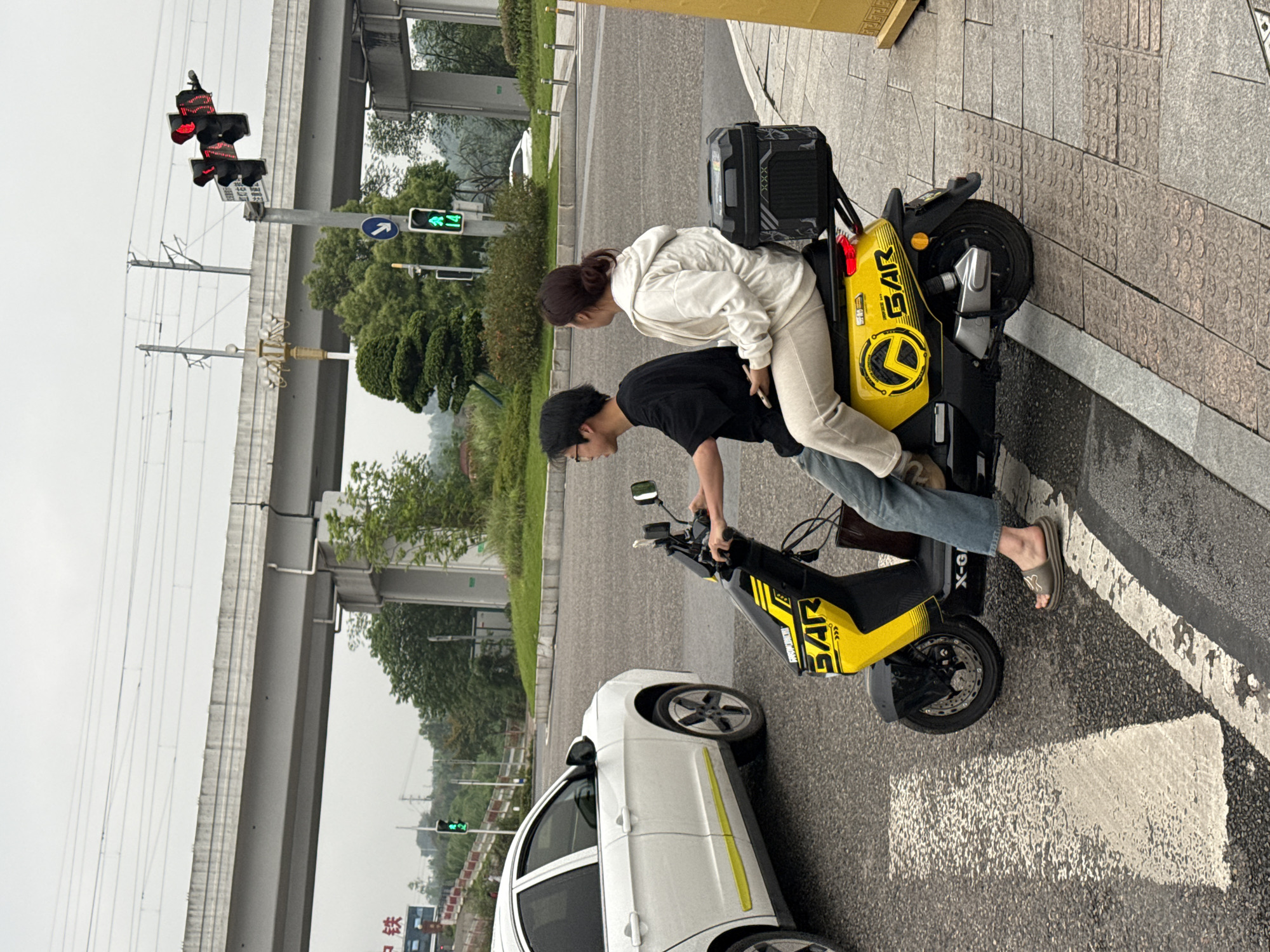Two people are riding a yellow and black electric scooter on a city street. A man is driving, wearing a black shirt, glasses, and a cap. A woman is riding behind him, wearing a white hoodie and beige pants. The scooter has a black cargo carrier attached to the back. A white car is parked nearby. A traffic light displaying a red light is visible overhead. The location is Jiangjin, China, as indicated by the Chinese characters visible in the lower left corner of the image.
