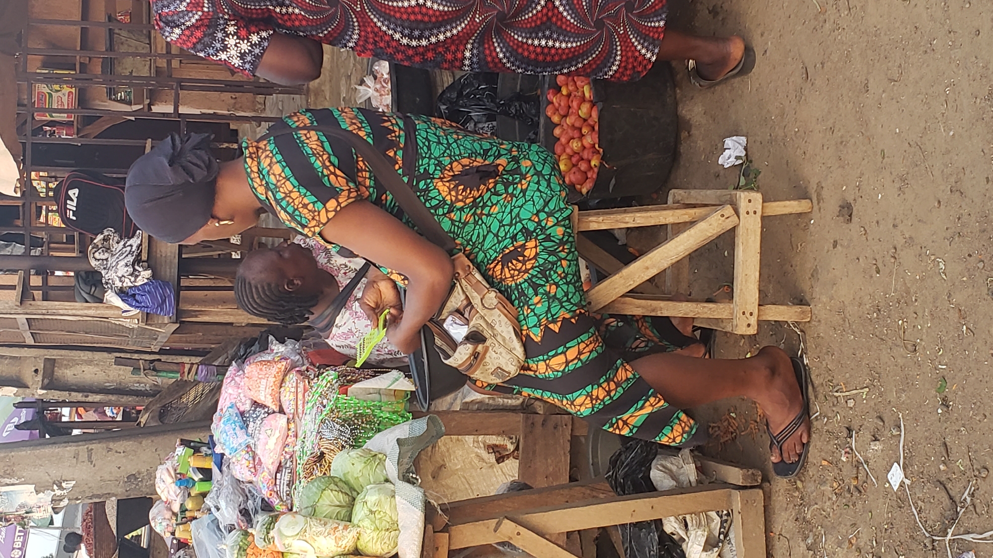 The image depicts a scene in Narayi, Nigeria. A woman wearing a green, orange, and black patterned dress and a black head covering sits on a small wooden stool. She has a patterned bag across her body and is looking down towards a second person, possibly a child, who is partially obscured. To her left are stacks of colorful items and produce, including cabbages. Behind her are tomatoes. The ground is dirt with scattered debris. Another person, partially visible, wears a patterned dress with red and black, and is standing nearby. A Fila bag is visible near the left edge of the scene.