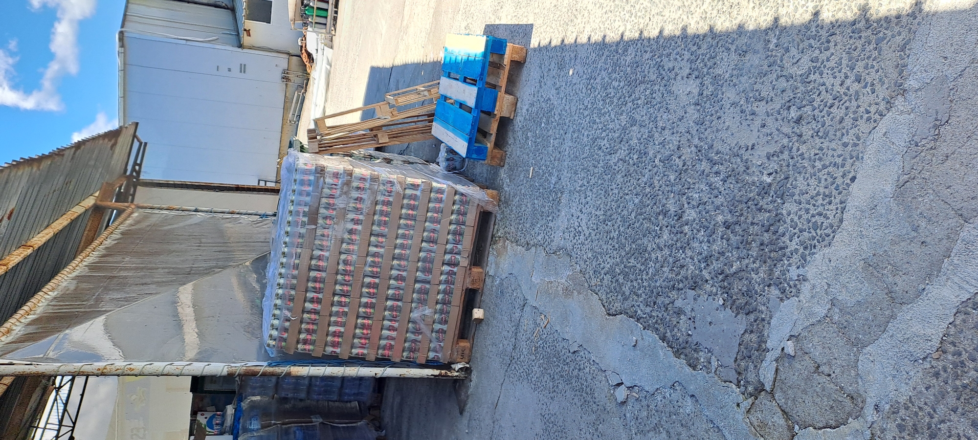 A pallet stacked with cans is leaning against a metal support beam, wrapped in plastic. The pallet is on a concrete surface, which is cracked and uneven, with small stones embedded in it. In the background, there is a large, white building with a corrugated metal roof and a cloudy blue sky above. To the right of the pallet, there is another pallet painted blue and yellow, and some wooden scaffolding. The scene appears to be an outdoor storage area, possibly in an industrial or commercial district, located in the city of Velika, Bulgaria.