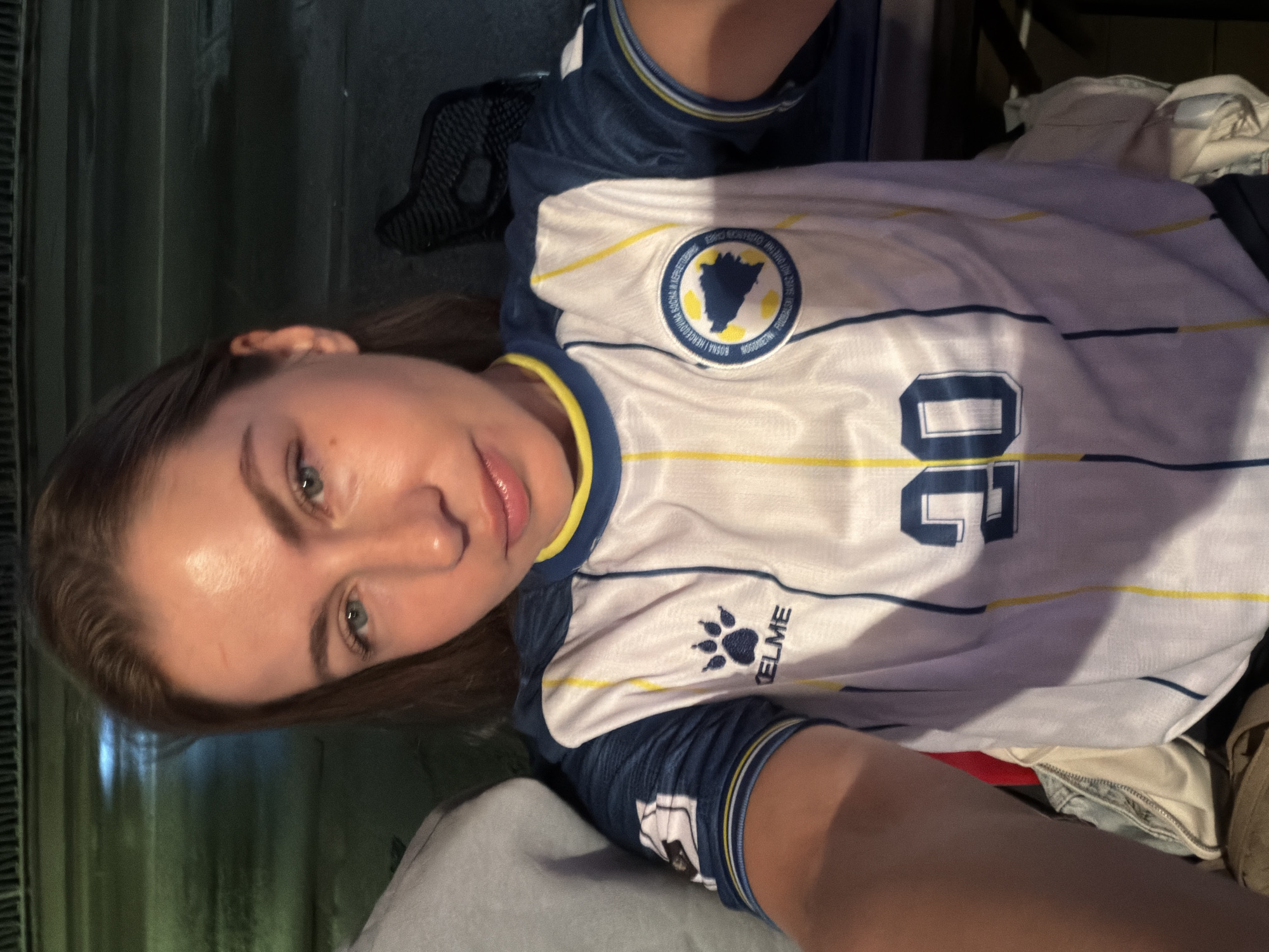 A young woman with blonde hair and blue eyes takes a selfie. She is wearing a white and blue soccer jersey with yellow trim and the number "20" in blue on the front. The jersey also features a crest with the outline of Bosnia and Herzegovina. The woman is outdoors at night, with a blurry background that suggests a city street. There are no explicit indications of the city of Travnik, Bosnia and Herzegovina within the image itself.