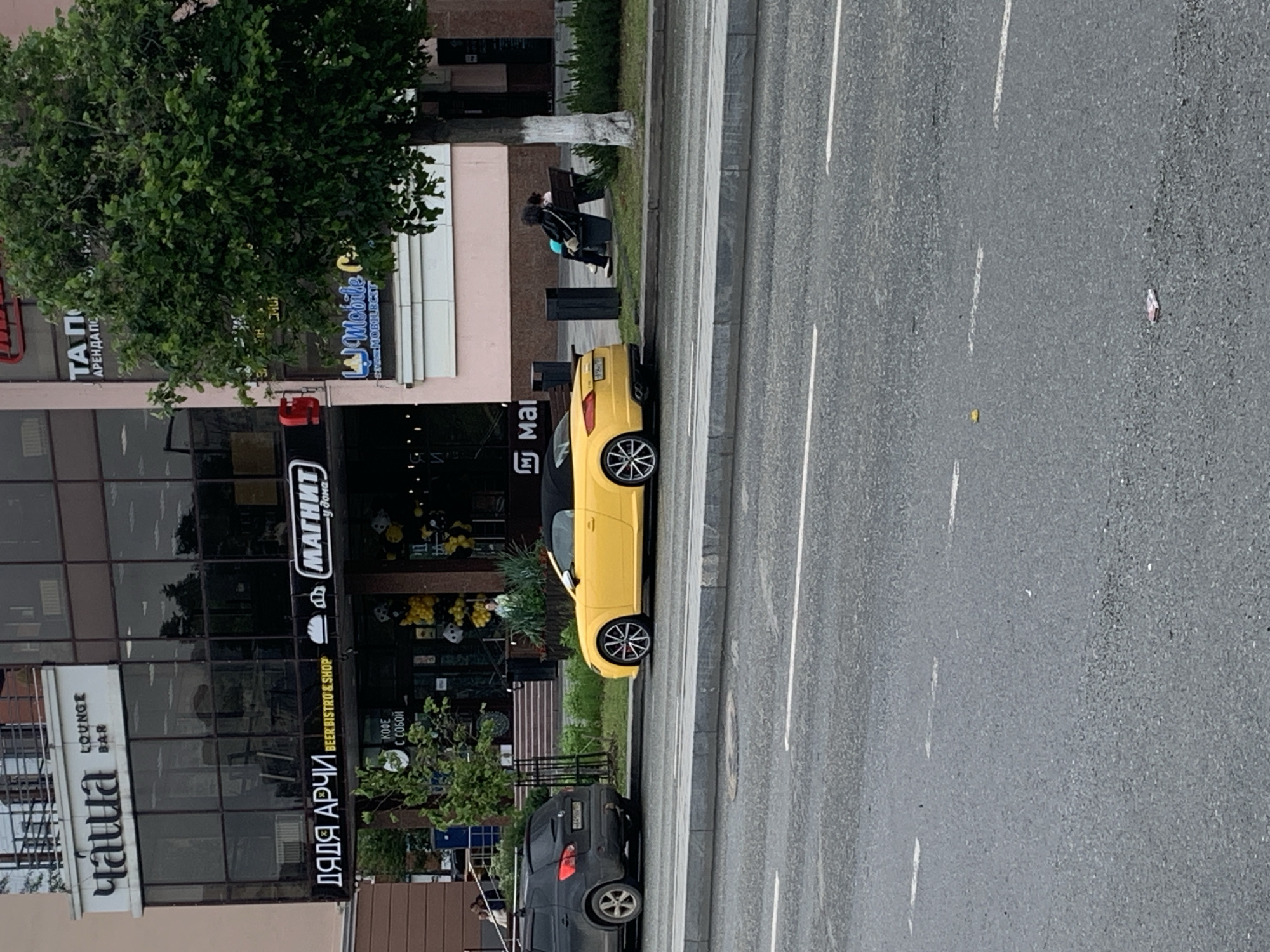In the city of Perm, Russia, on a cloudy day, a yellow convertible Audi TT is parked on the side of a street. The car is facing right, with its top down and black interior visible. The vehicle is situated in front of a multi-story building with a modern facade. The building houses several establishments, including a lounge bar named "Чаша" (Chasha) on the upper floor and a "Magnit" (Магнит) grocery store on the ground level. A dark gray SUV is parked to the left of the Audi.
To the right of the building, a large green tree stands by the sidewalk. A wooden bench is located beneath the tree, occupied by two individuals, one with dark curly hair and the other with lighter hair, facing away from the camera. In front of the building, there is a sidewalk area with several trash cans and some landscaping. The street in the foreground is paved and appears to be a multi-lane road with white lane markings. The overall scene suggests a commercial district within the city.