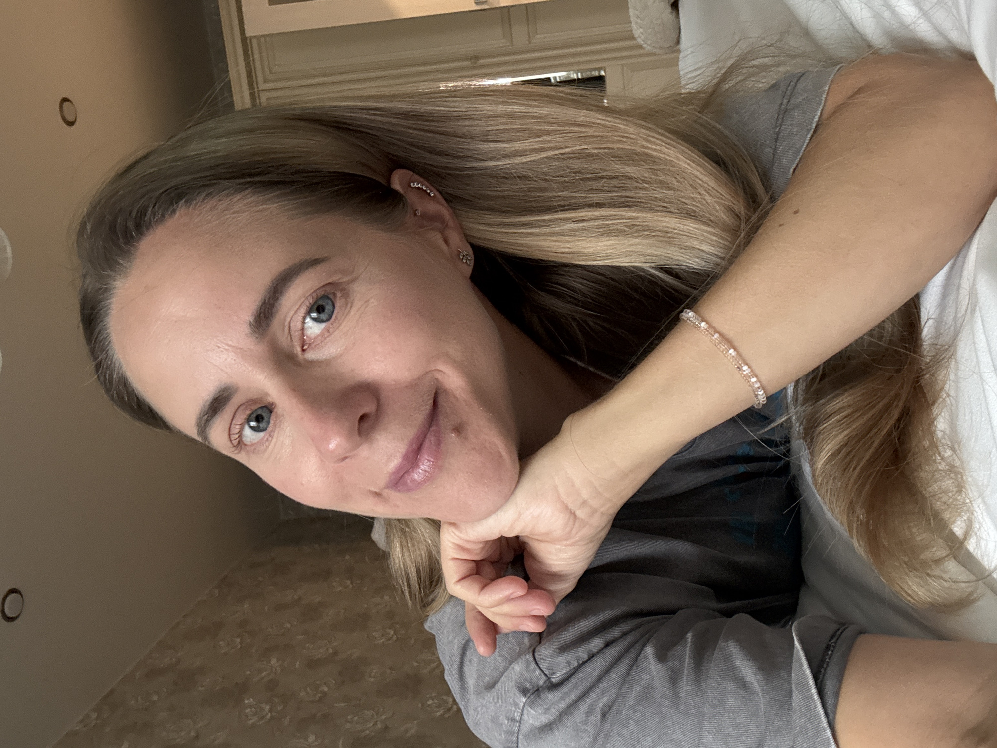 The image is a selfie taken indoors, likely in a bedroom, by a woman with blonde hair and blue eyes. She is wearing a gray t-shirt and a bracelet. Her hand is resting on her chin. The background shows a wall with patterned wallpaper and a closet. There is no indication of the location being in Nizhny Novgorod, Russia.