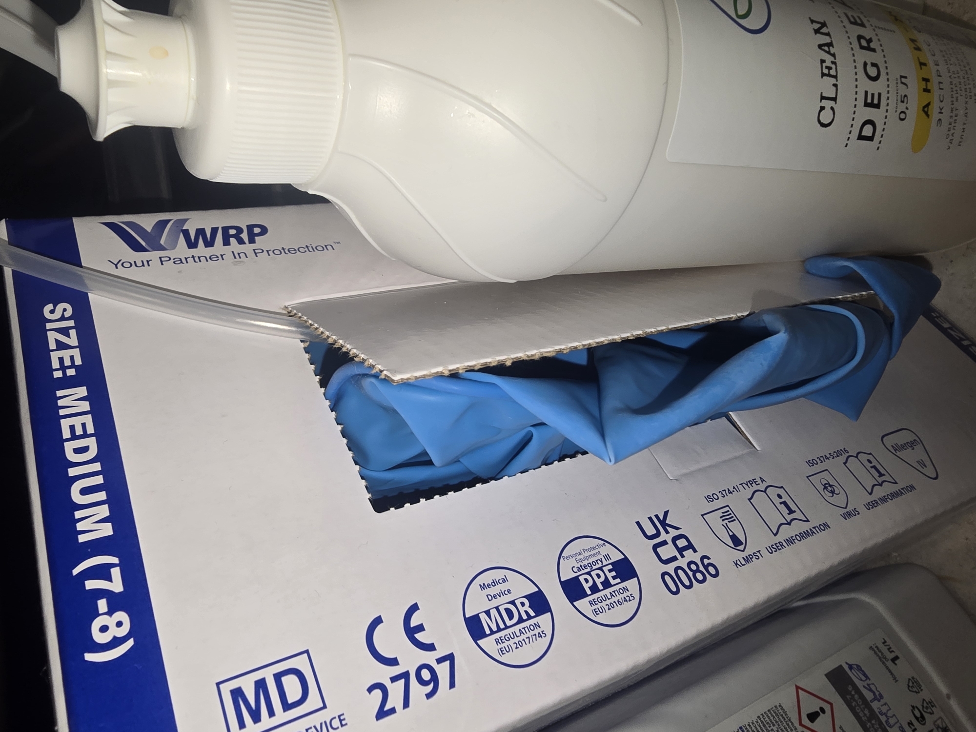 The image depicts a close-up of medical supplies, including a box of blue nitrile gloves and a white plastic bottle with a pump dispenser, likely containing disinfectant or hand sanitizer. The glove box features prominent labeling with "MD" and "CE 2797," indicating it is a medical device certified by the European Union. It also shows certifications for "MDR REGULATION (EU) 2017/745" and "PPE REGULATION (EU) 2016/425," along with other relevant symbols. The bottle's label is partially visible and reads "CLEAN DEGRES" and "0,5 л," suggesting a cleaning or degreasing agent with a volume of 0.5 liters. The background is dark, and the lighting suggests an indoor setting. There are no people or animals visible in the image. The scene appears to be a storage or display area for medical and hygiene products. There is no information to indicate the location is in the city of Ulan-Ude, Russia.