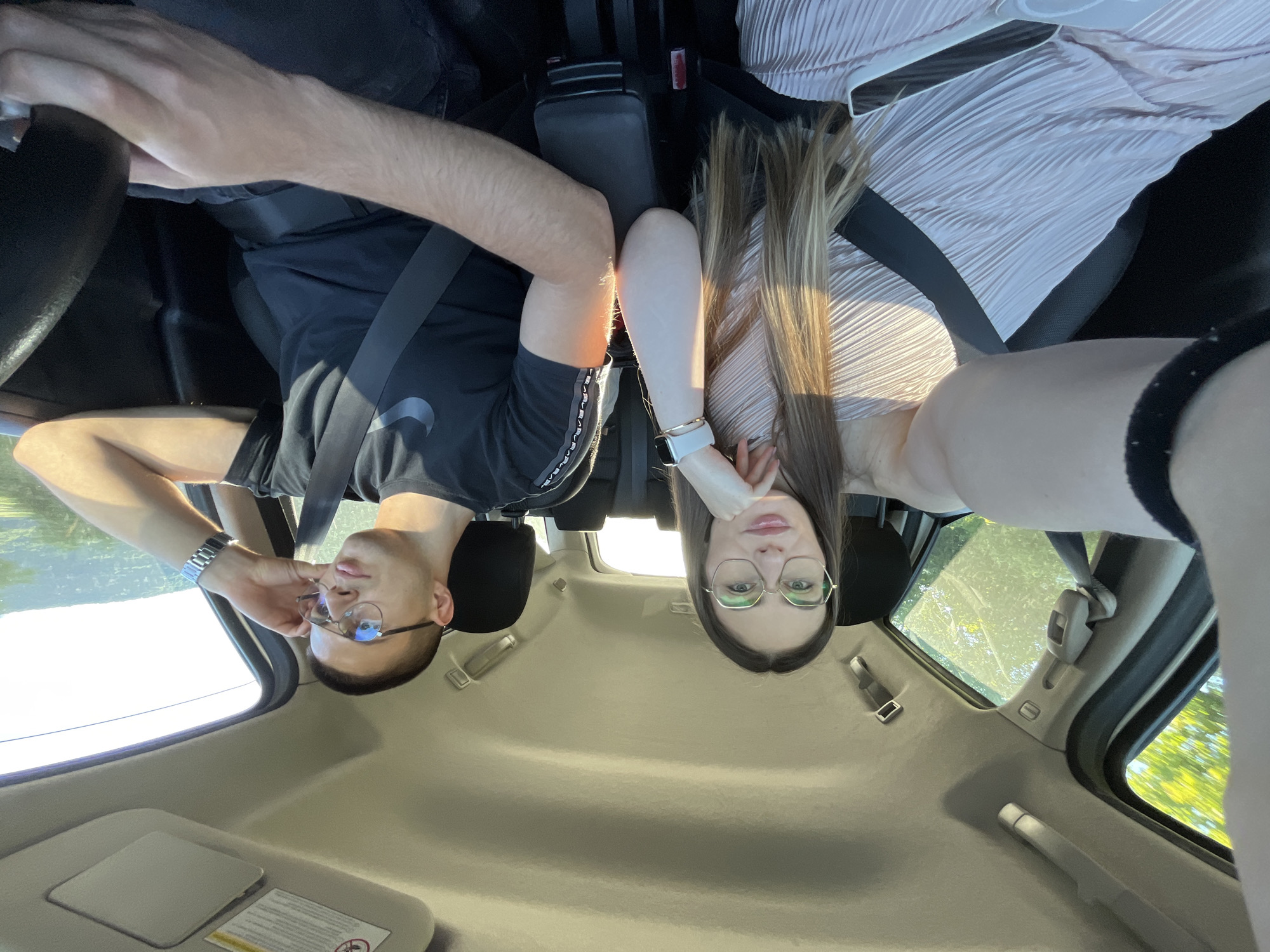 A medium shot from inside a car, oriented such that the top of the frame is the dashboard/roof. A man and a woman wearing eyeglasses sit in the front seats, both facing the camera. The man is on the left, wearing a black shirt with a white logo and a silver wristwatch. He has short brown hair and is wearing a seatbelt. The woman is on the right, wearing a light pink, pleated top and a silver Apple watch. Her long brown hair hangs downward. Both individuals appear to be looking at the camera. Outside the car windows, green trees are visible. The location is Unknown, Unknown.