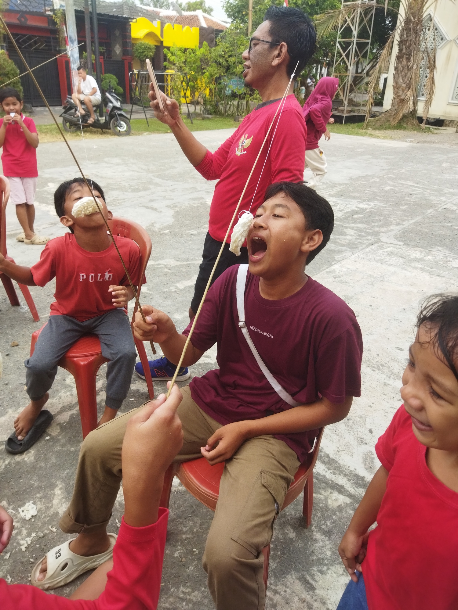 In an outdoor, paved area in Banjar, Indonesia, several children and adults are participating in a game. The focal point is a young boy sitting on a red plastic chair, attempting to eat a piece of puffed rice suspended by a string from a wooden stick held by an adult man. The boy's mouth is wide open, and the puffed rice is positioned in front of his face. Another young boy, also on a red plastic chair, is engaged in the same activity. A young girl in a red shirt stands in the background, holding a similar puffed rice snack. An adult man, wearing a red shirt with a national emblem, is standing and holding a mobile phone, appearing to film or photograph the game. A person in a pink hijab and beige pants is visible in the middle ground, walking away from the camera. The environment includes buildings with tiled roofs, some greenery, and a motorcycle parked in the background. The ground is made of rough concrete.