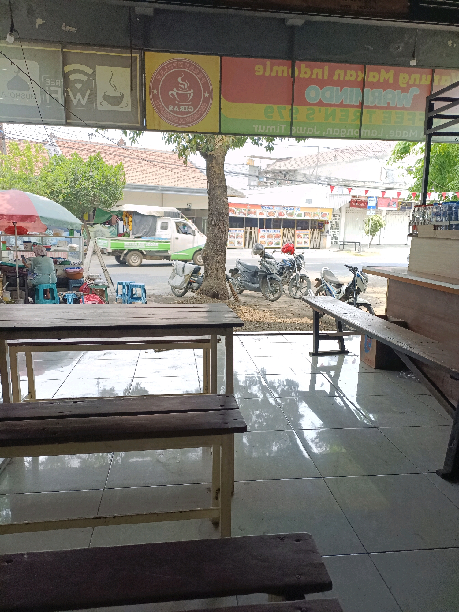 The image depicts a street-level view of a food stall or restaurant in Sukomulyo, Indonesia, looking outward from an indoor seating area. In the foreground, wooden tables and benches are visible, positioned on a tiled floor that reflects the overhead light and the scene outside. Through the open front of the establishment, the street scene unfolds.
To the left, a woman wearing a headscarf and a light-colored top is seated on a blue plastic stool at a food stall, appearing to be occupied with her phone. The stall itself has an umbrella providing shade, and various food items or ingredients are displayed. Next to the stall, a white and green truck is parked, with what appear to be fruits or vegetables in the back. Further down the street, several motorcycles are parked.
Above the street level, signs with text are mounted on the building. One sign, in yellow and red, displays text in what appears to be Indonesian. Another sign, in yellow with red text, shows a circular logo with a coffee cup and steam, alongside the words "PERKUMPULAN" and "GIRAS", likely indicating a coffee shop or cafe. Other signs show Wi-Fi symbols and a coffee cup icon. The background reveals buildings and trees, with power lines stretching across the scene. The overall impression is of a bustling, everyday commercial area in a tropical environment.