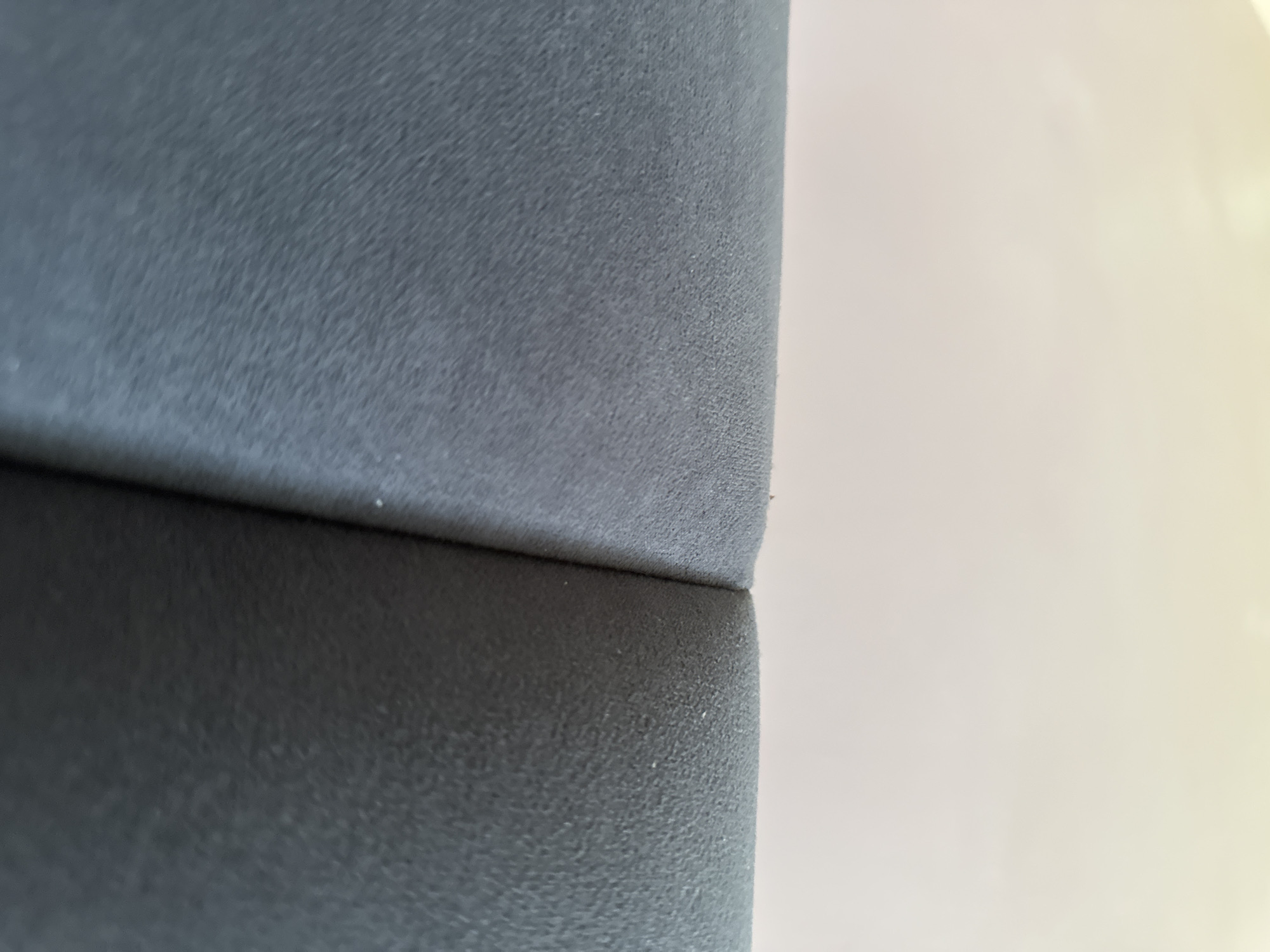 A very close-up, angled shot of a dark gray fabric surface with a subtle texture of fine bumps. The fabric appears to be part of a soft furnishing, possibly upholstery. The surface is divided by a slight seam down the center, creating two rounded edges that meet. The background is a soft, out-of-focus blur of light beige. The lighting is diffused, casting no harsh shadows. The scene is devoid of any discernable urban context or specific location.