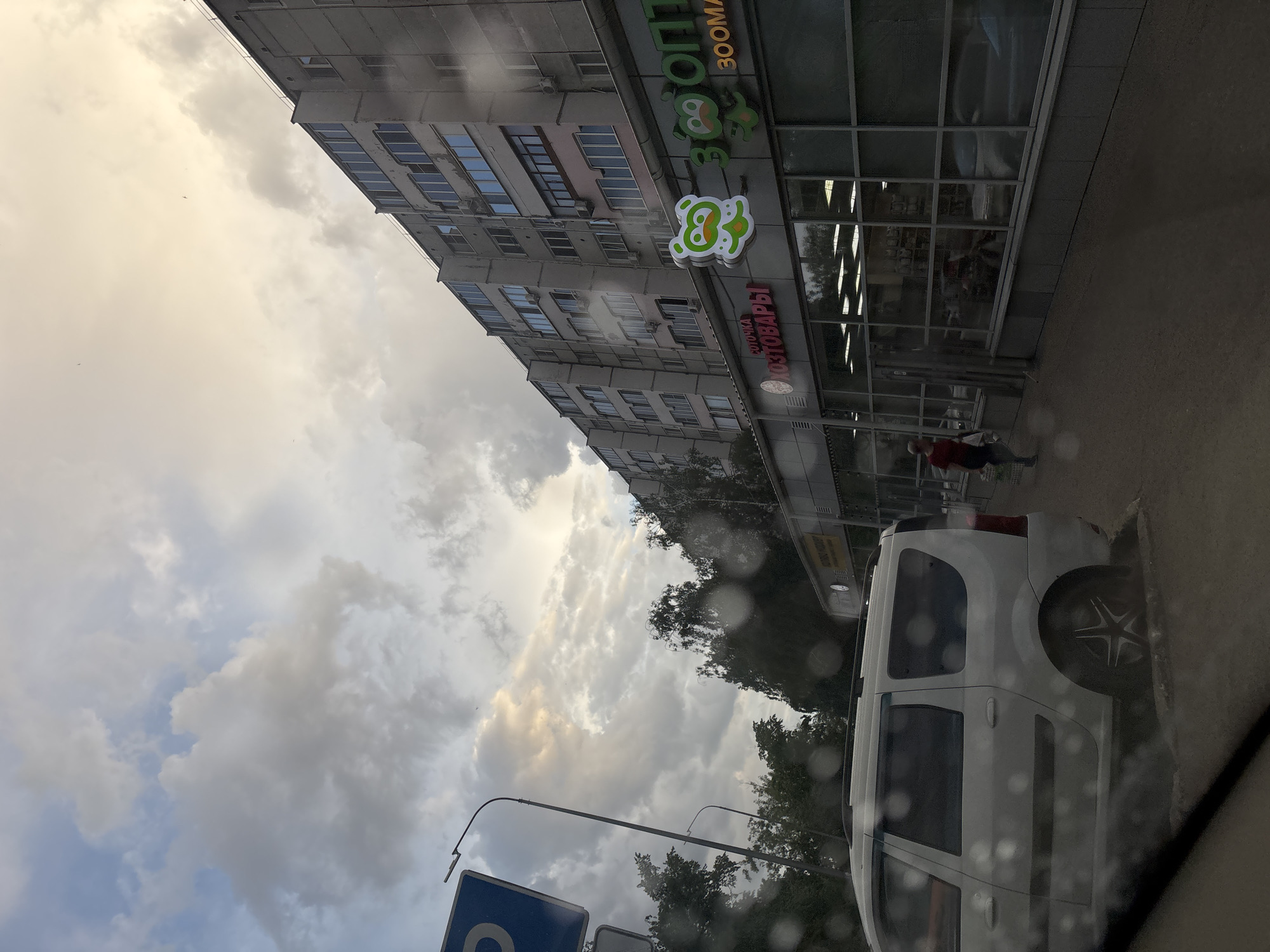 The scene is a street view in an urban environment, likely observed through a vehicle window due to raindrops visible on the glass. Dominating the right side of the frame is a white van parked on the curb, positioned diagonally from the lower right corner towards the upper center. A multi-story building, characterized by repetitive rows of windows, extends upwards and slightly to the left of the frame.
Located on the building's facade is a commercial establishment identified by illuminated signage in Cyrillic script, with a prominent stylized animal logo. A person is walking along the sidewalk in front of the store.
In the lower left corner of the frame, there are road signs mounted on poles. The sky overhead features a cloudy atmosphere. Streetlights are visible among the trees.