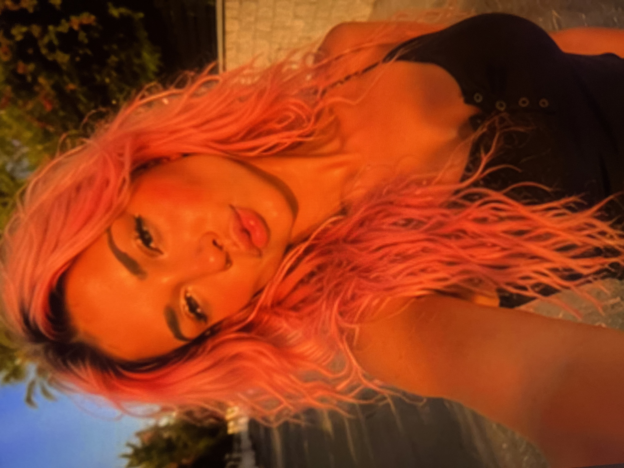 A woman with vibrant orange hair is captured in a selfie, her face turned towards the camera with her lips pursed. She is wearing a dark, buttoned tank top. The background suggests an outdoor setting with blurred foliage and a wall, possibly in a residential area. The lighting indicates it might be sunset or sunrise, casting a warm, orange glow over the scene.