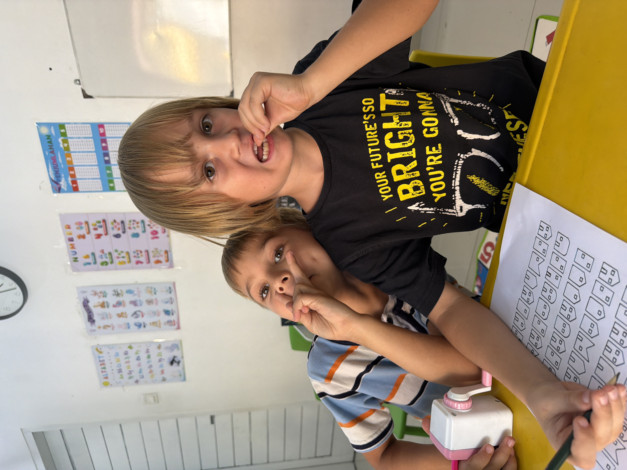Two young boys are seated at a bright yellow table in what appears to be a classroom or study area in Kuta, Indonesia.
The boy in the foreground, with light brown hair, is wearing a black t-shirt. He looks directly at the camera with wide, expressive eyes, his mouth slightly open, and his fingers near his teeth in a playful gesture. His t-shirt prominently displays yellow text that reads, "YOUR FUTURE'S SO BRIGHT YOU'RE GONNA" (with the last word partially visible).
Behind him, another boy with darker hair, wearing a blue, white, and orange striped t-shirt, is also looking towards the camera, though his gaze is less direct. He has one hand near his face and is holding a dark pencil in his other hand, poised over a piece of white paper on the table. The paper features repetitive outlines of small house-like shapes, suggesting a tracing or drawing activity. A white and pink manual pencil sharpener is also on the table.
The background consists of a white wall adorned with several educational posters. These include a poster titled "PENJUMLAHAN" (Indonesian for "Addition"), another for "NUMBER," and an "ALFABET" (Alphabet) chart. A whiteboard and a wall clock are also visible on the wall. The overall scene captures the children engaged in their activities with lively expressions.