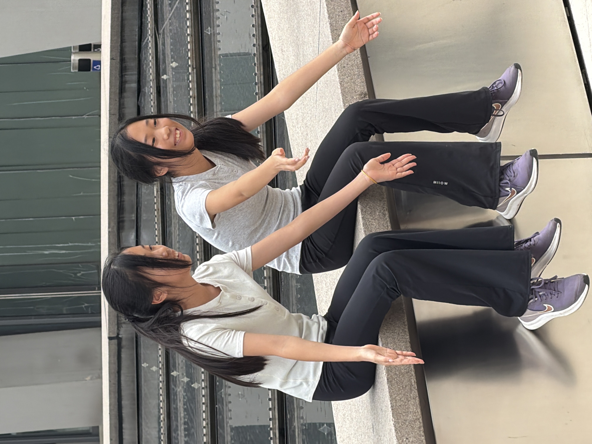 Two young Asian females are sitting on a ledge outdoors. Both have long, dark hair and are wearing dark pants and light-colored t-shirts. They are positioned side-by-side, facing left, with their legs dangling over the edge of the ledge. The female on the left is wearing a light gray t-shirt with buttons at the neckline and dark pants. Her right arm is extended forward, with her hand open. The female on the right is wearing a light gray t-shirt and dark pants with the word "MIIOW" visible. Her arms are both extended forward, with her hands open. Both are wearing purple athletic shoes with white soles and Nike swoosh logos. The background shows tiered steps and a metallic wall. The scene appears to be in an urban environment in Yuzhong District, China.