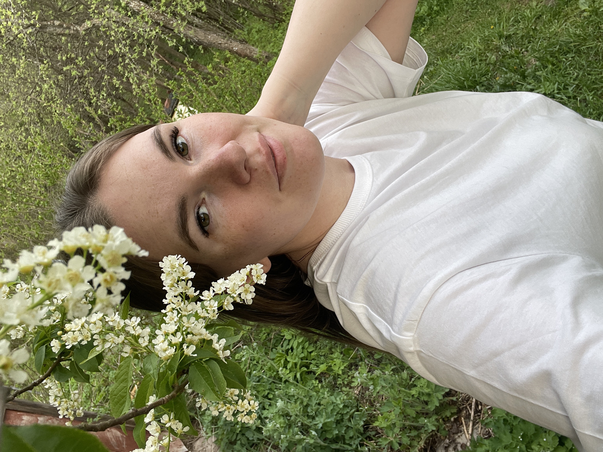A fair-skinned woman with brown hair lies on green grass, facing the camera. She wears a white t-shirt and has her arms raised behind her head. The foreground features a flowering branch with white blossoms and green leaves. The background consists of green foliage and trees. The location is Zhigalki, Russia.