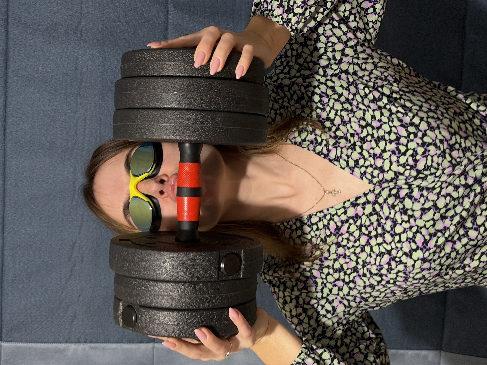 A woman with blonde hair, wearing yellow and green sports goggles, is lying on a blue surface, possibly a couch or bed. She is holding a dumbbell above her face. The dumbbell is composed of multiple black weight plates, secured by a red and black handle. The woman is wearing a patterned dress, with hues of green, purple, and black. A silver necklace is visible around her neck. Her fingernails are painted pink. The photograph appears to have been taken indoors and rotated. The location is not evident from the image.