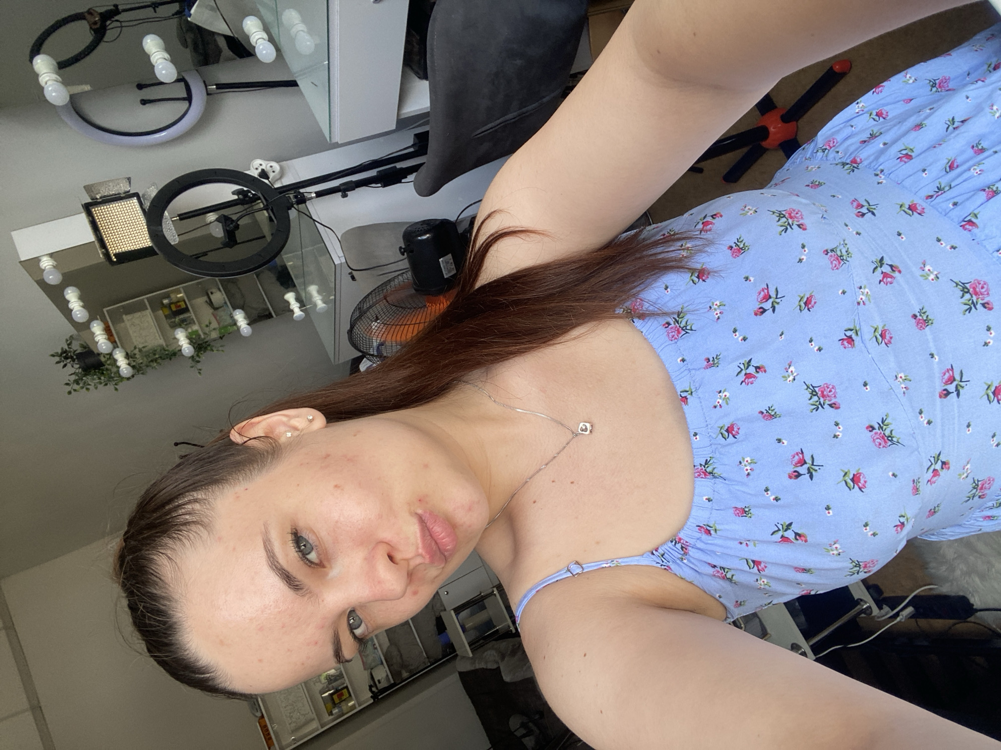 A young woman with auburn hair tied in a high ponytail and blue eyes is shown from the chest up, taking a selfie in an indoor setting. She is wearing a light blue sundress with a floral pattern of pink roses and white flowers. She has a silver necklace with a small square pendant. Her facial skin shows some blemishes. In the background, there are shelves stocked with various items, a makeup vanity with ring lights and a professional camera light, and a black office chair. The environment suggests a personal workspace or studio. The image was taken in Poltava, Ukraine.