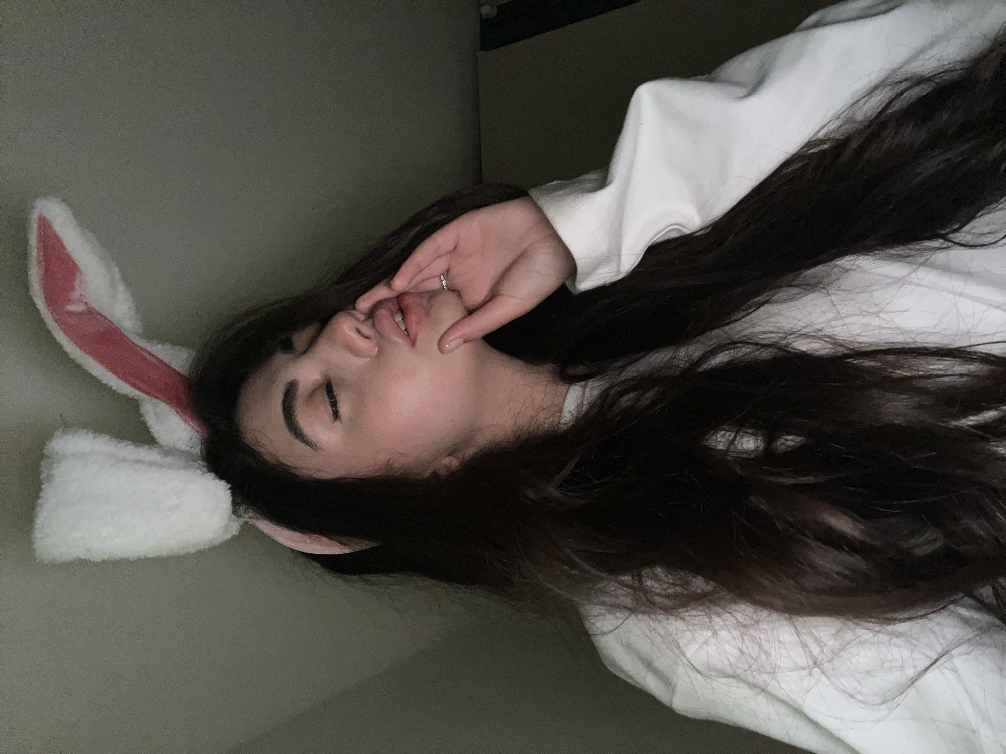A young woman with dark, long hair and white bunny ears on her head is captured in a close-up, dimly lit selfie. She is wearing a white sweatshirt. Her eyes are closed, and her lips are pursed as if she is blowing a kiss or whispering. Her right hand is raised to her face, with her fingers gently touching her lips and cheek. A silver ring is visible on her ring finger. The background is a muted grey wall, indicating an indoor setting, possibly a bedroom or living space. The lighting is low, creating shadows and highlighting her face and the texture of her hair and ears. The overall mood is playful and intimate. There is no visible indication of the location being in Chotěšov, Czechia.