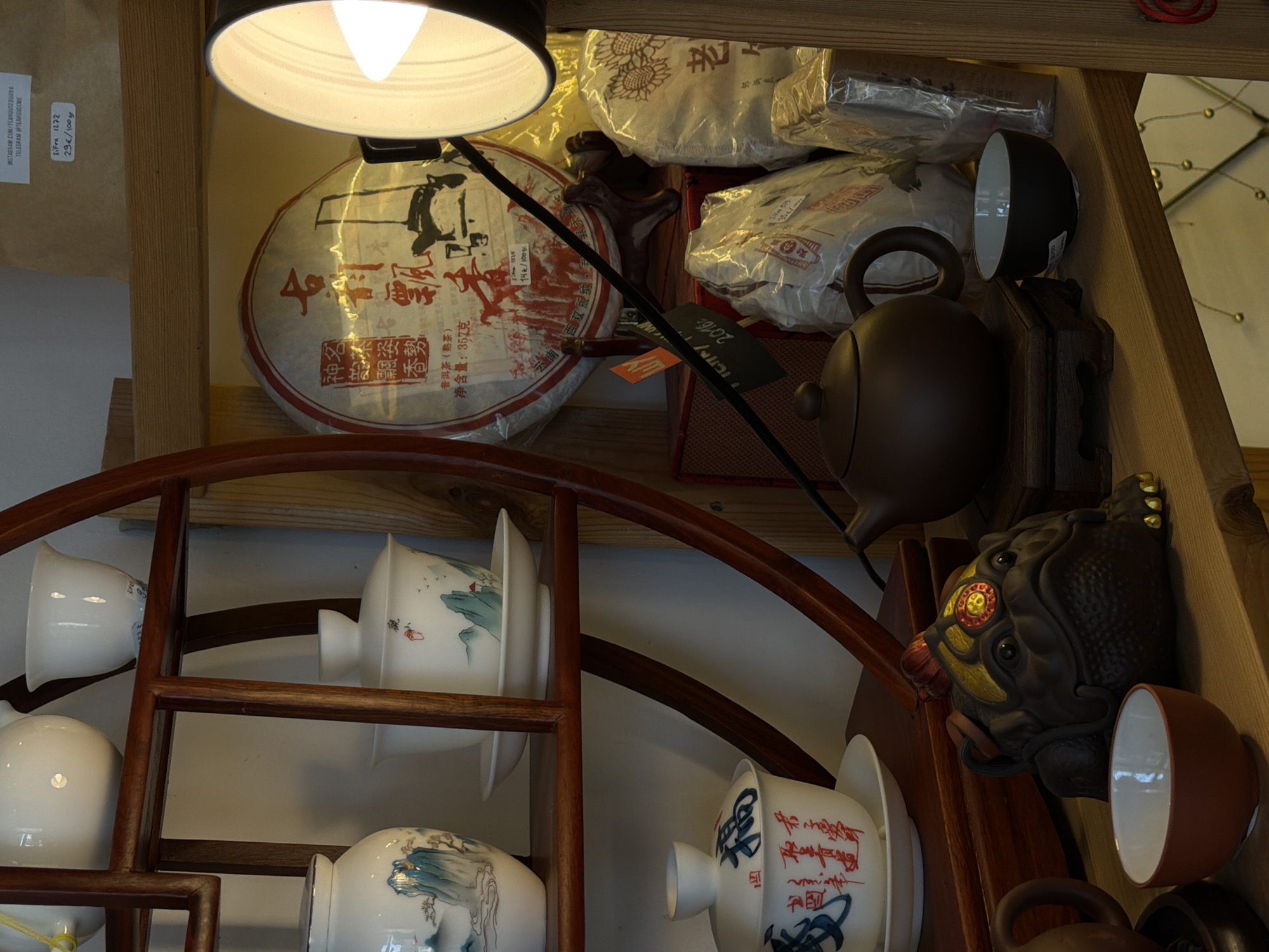 The image shows a shelf full of tea-related items. There is a light illuminating a round, flat cake of what appears to be tea. Text in Chinese is visible on the tea cake. Behind it, there are packages of tea. A brown teapot is also visible, as well as a decorative frog-like object with gold accents. Below the shelf, there is a wooden display with teacups. The OCR text indicates this is from a tea house in Budva, Montenegro. Specifically, information in the text refers to instagram.com/tearousebudva and telegram @teahouseme. There is pricing information for tea on a label: Sifre 1272, 29€/100 gr. The tea cake is labeled 普洱茶(熟茶), meaning Pu-erh tea, and contains a net weight 净含量:357克. There is a note that reads 南西双版纳, indicating it is from Xishuangbanna, Yunnan. The tea cake is labeled 2016, indicating it is from 2016.