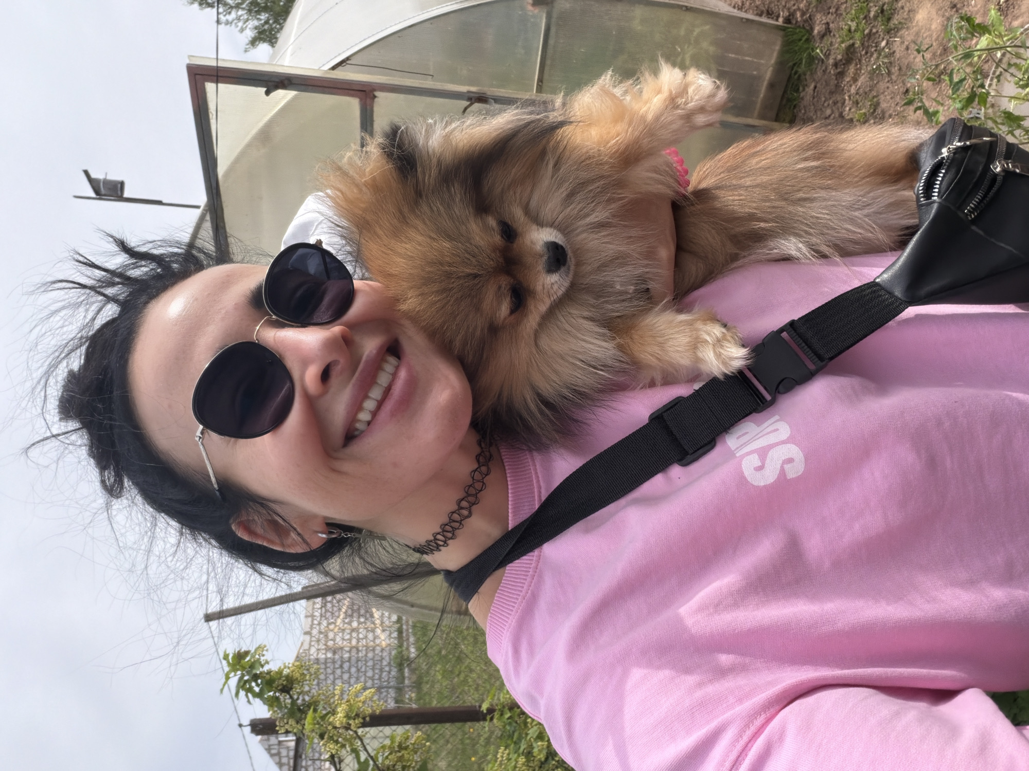 A woman is holding a Pomeranian dog on her shoulder. She is wearing a pink sweatshirt with a white "SR" on the sleeve, round sunglasses, and a black choker. She also has a black fanny pack across her chest. The dog is mostly brown, but has a black nose. The background includes a greenhouse, trees, and a cloudy sky. The location is in the city of Бронница, Russia.