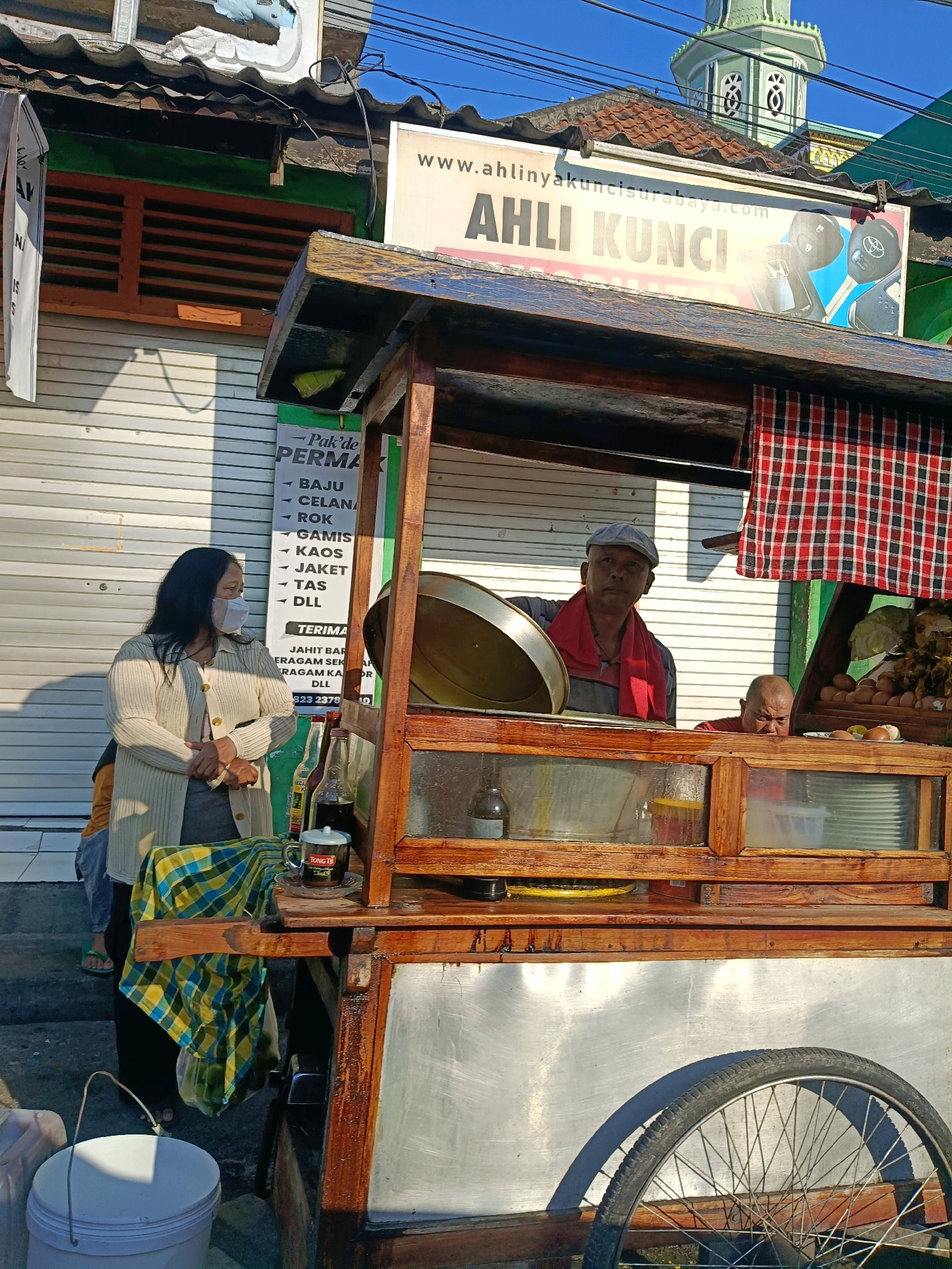 This is a street scene in Surabaya, Indonesia, featuring a food cart. A woman wearing a white mask and a cream-colored cardigan stands to the left of the cart, facing right. Behind her, a sign with "Pak'de PERMA" and a list of clothing items is visible.
The food cart is made of wood and metal. A man wearing a grey cap and a red scarf is standing behind the counter of the cart, looking towards the viewer. To his right, another man with a bald head, wearing a red shirt, is peering out from the cart. The cart displays various items including eggs, food, and bottles of liquid. A red and black checkered cloth is draped over the cart.
In the background, a building with a green roof and a minaret is visible, suggesting a mosque. The overall setting appears to be a sunny day on a street. The building behind the cart has a shuttered window and a sign that reads "AHLI KUNCI SURABAYA.COM AHLI KUNCI". A bicycle wheel is attached to the side of the cart.