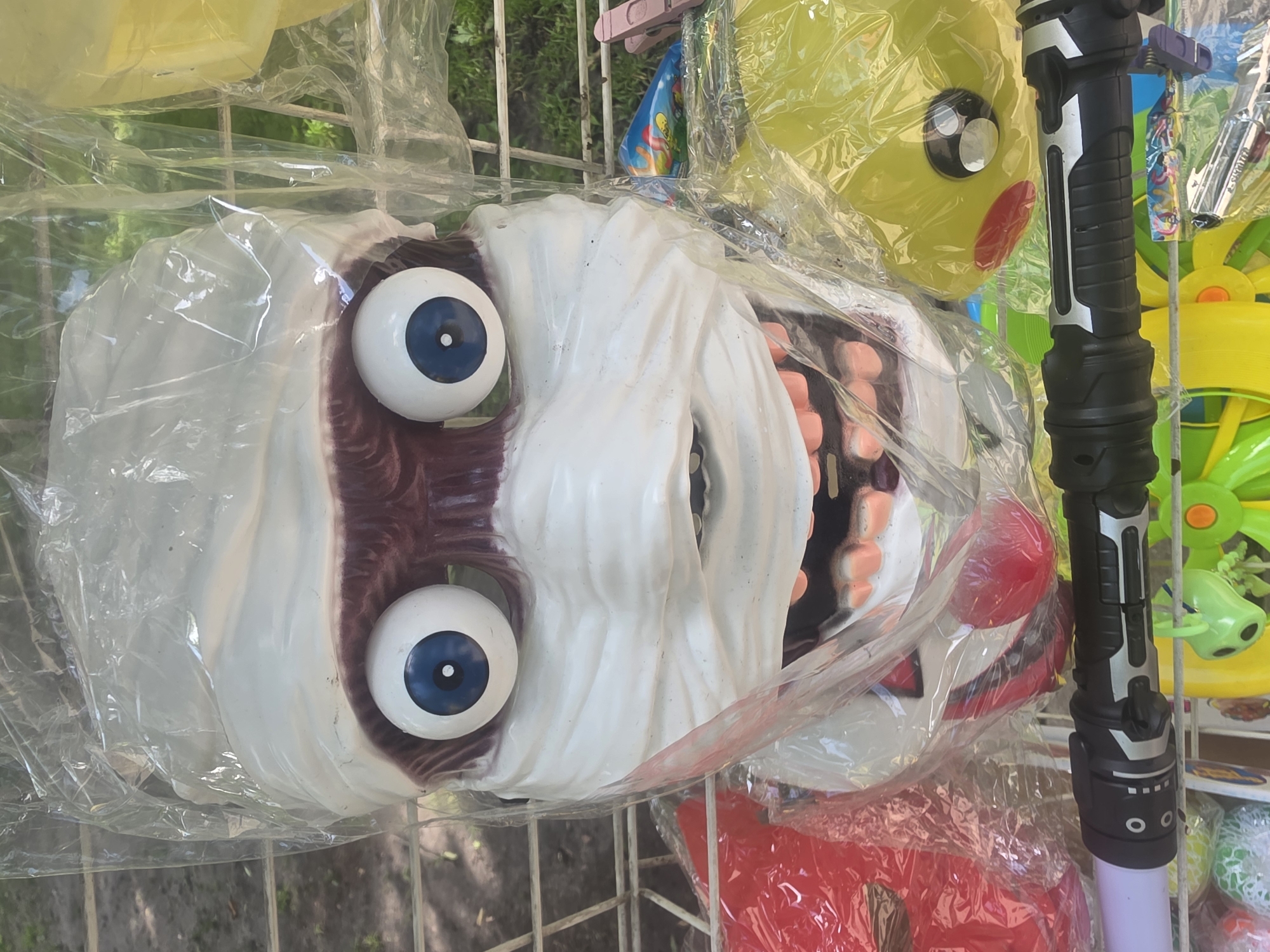 The image displays a close-up, eye-level view of a plastic mummy mask, packaged in clear plastic, hanging outdoors. The mask is white with large, wide-set blue eyes and a gaping mouth revealing rows of teeth. It is wrapped with white bandages, giving it a mummified appearance. The background is slightly blurred, showing a grid structure, foliage, and other colorful items, suggesting it's a display for sale. The scene appears to be outdoors, possibly in a market or store. The image was taken on the street of Bila Tserkva, Ukraine.