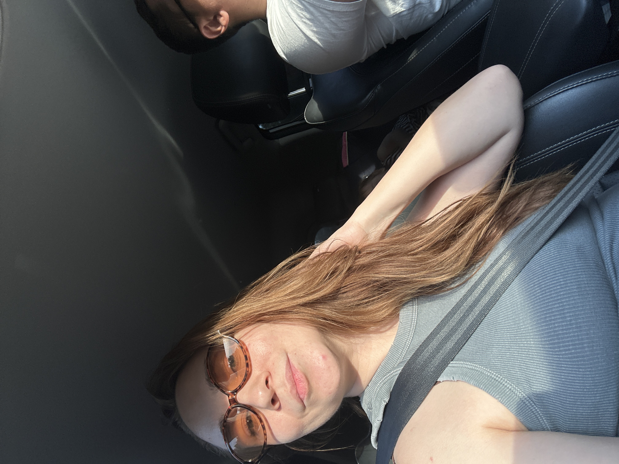The image shows a woman with reddish-brown hair wearing sunglasses and a seatbelt, presumably in a car. Another person, a man, is visible in the passenger seat in the background. The car's interior is visible, including the black leather seats. The setting appears to be outdoors, based on the natural lighting. The location in Докша, Russia, is not directly verifiable from the image itself.