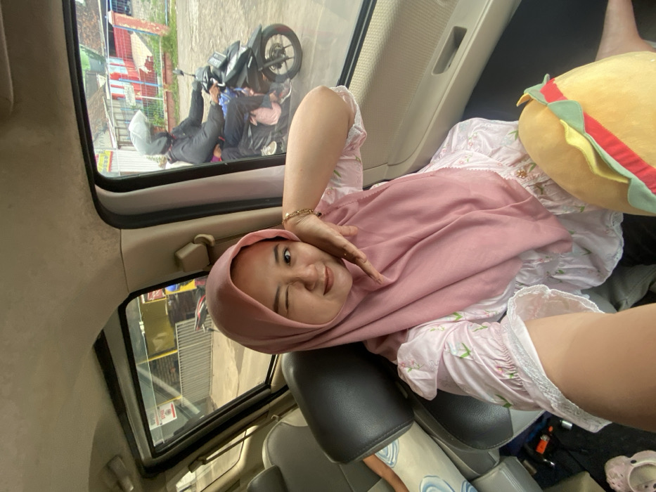A person wearing a pink hijab and a floral-patterned dress poses inside a car, winking at the camera. A plush burger toy is visible next to them. Outside the car window, a street scene includes a motorcycle with two people nearby, one lying on the ground and another sitting on the bike. The car interior shows gray seats and a window.