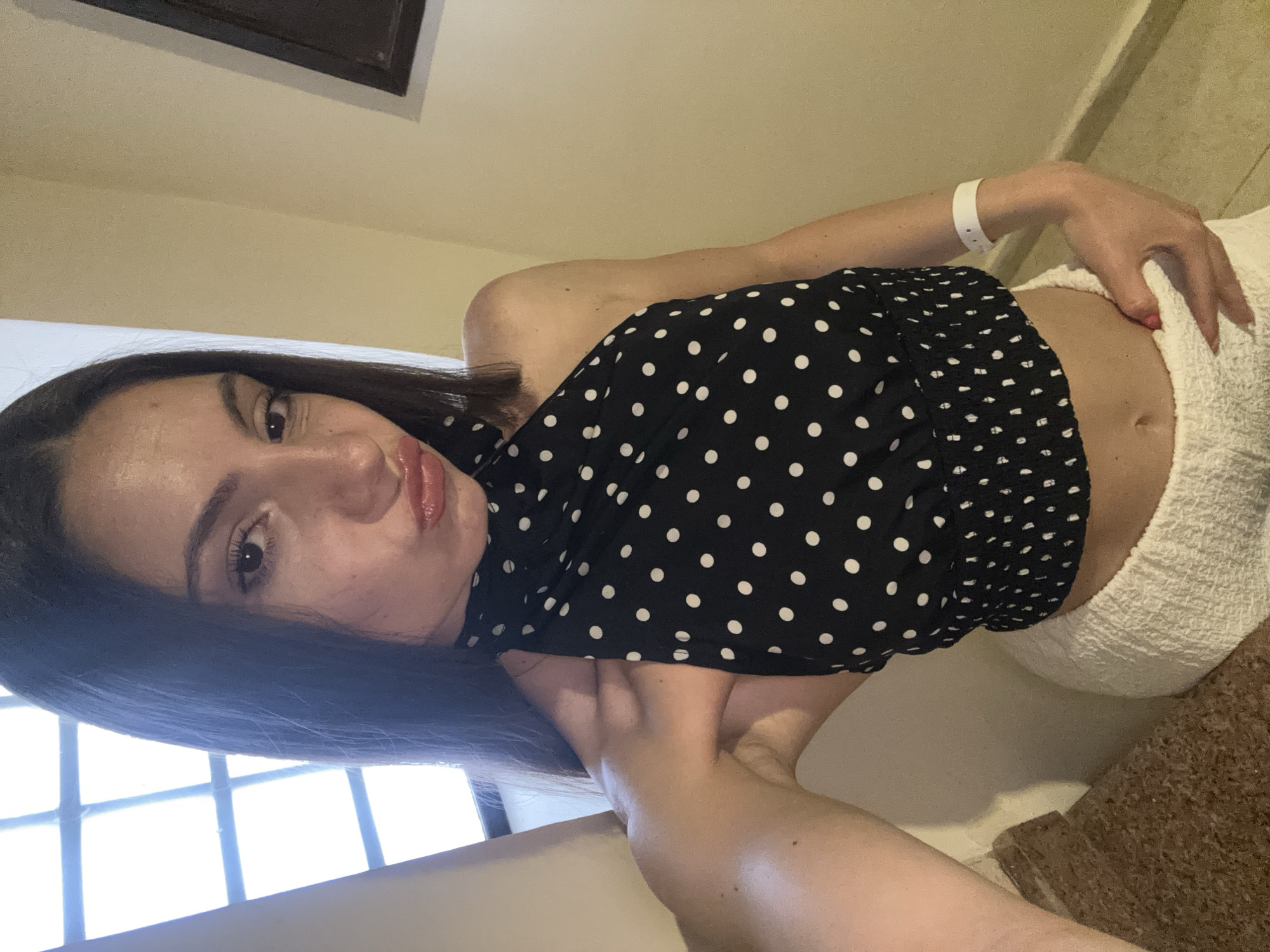 A woman is posing indoors. She is wearing a black polka dot top and white shorts. She has long dark hair and is positioned near a window. She is wearing a white hospital bracelet.
