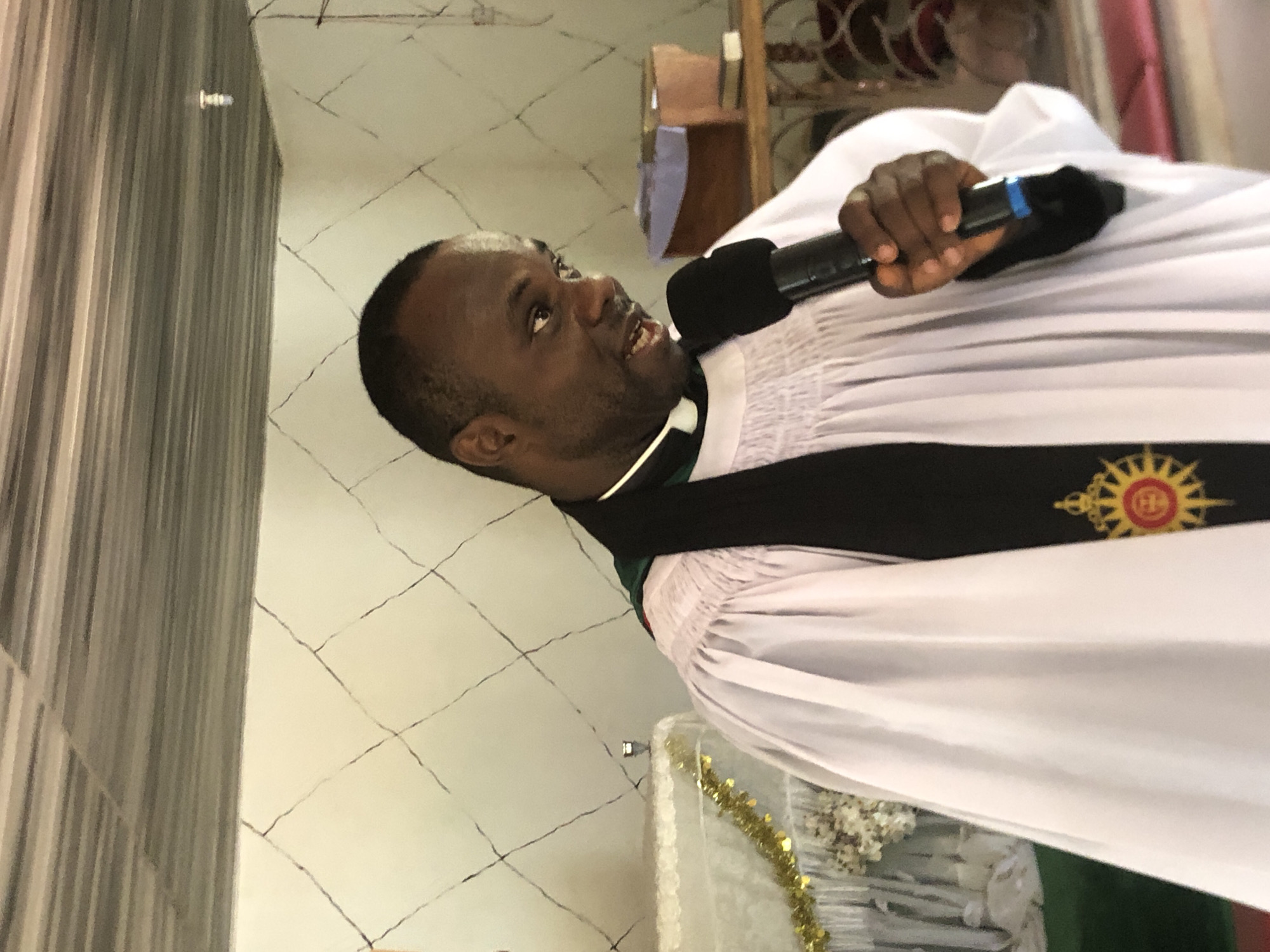 A medium, eye-level indoor shot depicts a dark-skinned male in a white robe with a black stole. He's holding a black microphone close to his mouth and appears to be speaking. The scene is in Ukwu-Abwa, Nigeria. The walls show a cracked tile pattern and there's a curtain on the left. In the background is a shelf and a table decorated with gold tinsel and white flowers. The overall lighting appears bright.