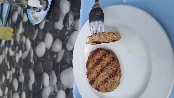 A close-up photograph, taken from an elevated perspective, captures a meal served on a table in Ezerovo, Bulgaria. The central subject is a grilled, round meat patty, approximately 10-12 cm in diameter, positioned on a white ceramic plate. The patty displays distinct dark brown grill marks and a coarse texture with visible green specks, indicating herbs. A silver-colored metal fork, inserted into the patty, lifts a portion revealing the uniformly cooked, light brown interior.
The white plate rests on a light blue plastic tray. The table surface beneath features a distinctive pattern resembling an arrangement of smooth, rounded white and dark gray/black stones or pebbles. In the upper right background, a small, circular plate with a multi-colored floral pattern holds a metal condiment caddy containing two shakers. Further to the left, a clear plastic bottle, partially filled with a pale yellow liquid, is visible. No individuals are present within the frame.