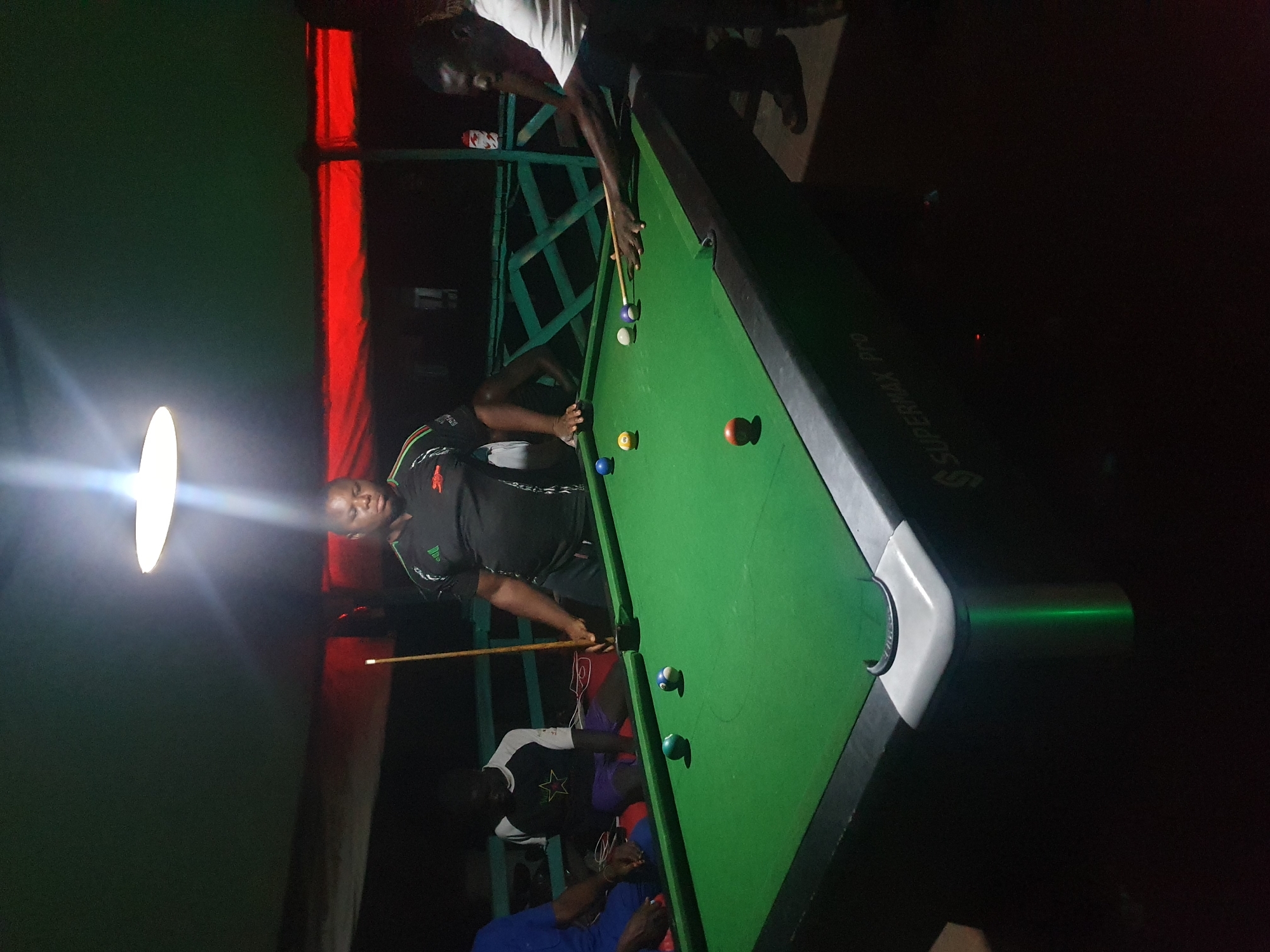 A group of men are playing pool at night, under a dimly lit ceiling fan. The pool table is green with white corner pockets. Several balls are scattered across the table, including a solid red ball, a striped yellow ball, and a solid blue ball. One man, wearing a black and green t-shirt, stands over the table with a cue stick, preparing to take a shot. Another man, in a white t-shirt, is also by the table, watching the game. Two other men are seated in the background, one in a blue shirt and the other in a white and black shirt. The setting appears to be an outdoor or semi-outdoor establishment in Lokoja, Nigeria, given the presence of a red tarp and a wooden railing in the background, suggesting a rustic and informal environment. The lighting is minimal, casting long shadows and highlighting the pool table.
