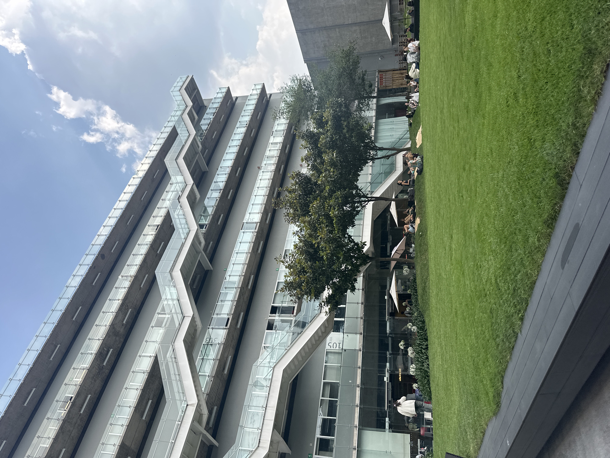 The image depicts a modern, multi-story building in Mexico City, Mexico, viewed from a low angle. The building features a unique architectural design with prominent external staircases zig-zagging up its facade. The exterior consists of gray concrete or similar material, accented with glass panels along the stairways and windows. In front of the building is a green lawn area where people are sitting and relaxing. There are trees and white umbrellas along the lawn, providing shade for the individuals present. Some people are standing near the base of the building. The sky is visible above the building with a mix of blue and white clouds. A portion of another building appears in the top right of the image.