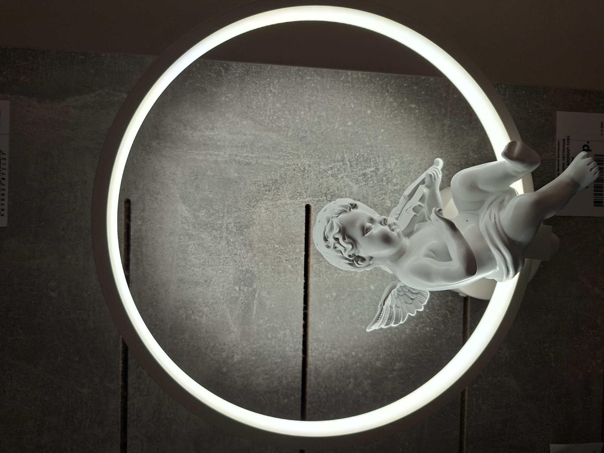 A white porcelain statue of a cherub playing a violin is illuminated by a circular LED light. The statue is positioned in the center of the circular light, which is mounted on a dark gray, textured wall. The cherub is seated with its legs crossed, and its wings are spread. The light casts a soft glow on the cherub and the surrounding wall. A white label with Cyrillic text is visible below the statue, attached to the wall. The scene appears to be an indoor setting, possibly a home or a shop display. There is no information in the image to suggest a specific location within the city of Izhevsk, Russia.