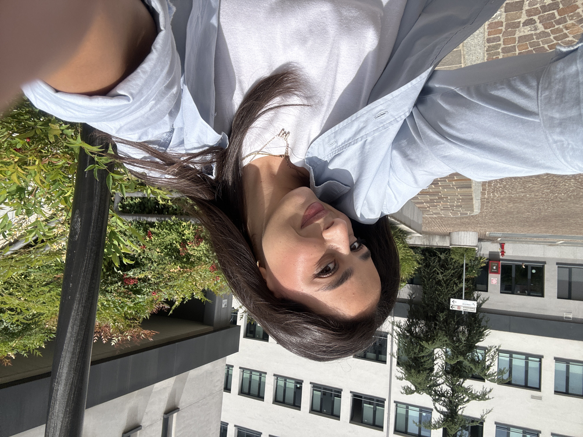 A young woman takes a sunlit outdoor selfie, her head tilted slightly and her dark hair falling across her shoulder and chest. She has fair skin, dark eyes, and a neutral-to-softly smiling expression. She wears a light blue button-up shirt open over a white t-shirt, with a delicate gold-toned necklace featuring a small "M" or "W" charm visible.
The scene is an urban environment, likely in Milan, Italy, given the discernible text. Bright sunlight casts clear shadows, indicating it is daytime. In the background, modern, multi-story buildings with numerous dark-framed windows are visible. Lush green foliage with red berries grows alongside what appears to be a dark vertical pole, suggesting an elevated vantage point like a balcony or terrace.
A street sign, partially obscured and appearing upside down due to the image's orientation, shows an arrow pointing left and another pointing right. The text on the right-pointing arrow reads "VANITA" on the top line, "UNIONE EDILIZIA" on the middle line, and "COSTR. VANTIA" on the bottom line. A small tree stands in front of the buildings, and a red fire alarm box is mounted on one of the grey building walls. The ground in some cropped areas shows a patterned cobblestone or brick surface.