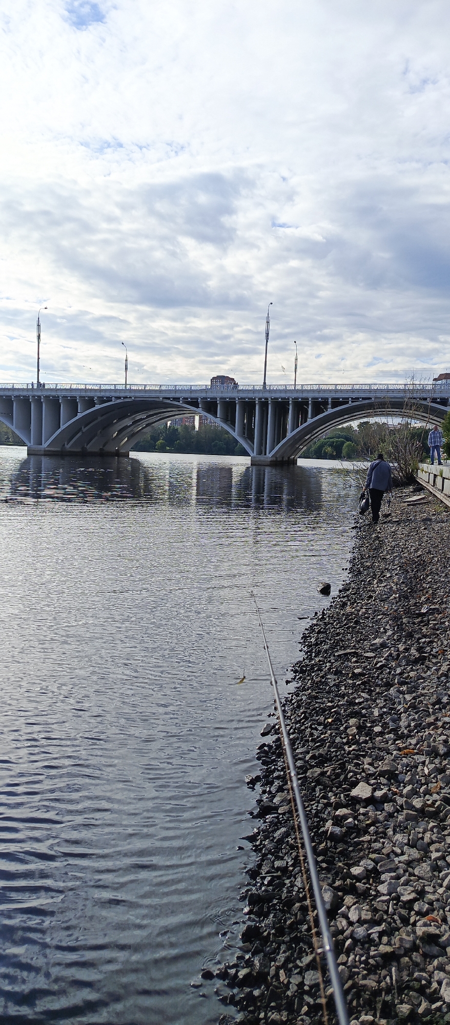 An overcast day in Yekaterinburg, Russia, features a wide, tranquil river with a modern, concrete bridge spanning its width. The bridge has multiple arched sections and is illuminated by streetlights. The riverbank on the right side of the image is lined with rocks and a few sparse bushes. Two individuals are present: one person is walking away from the camera along the riverbank carrying a dark bag, and another person, wearing a plaid shirt, stands further down the bank. A fishing rod is positioned in the foreground, extending into the water, with a small lure visible. The background shows a treeline and some buildings beyond the bridge.